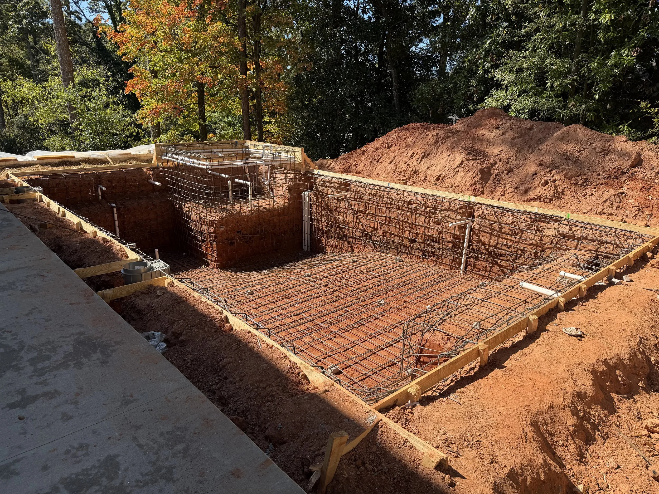 Construction site with exposed red soil, wooden framing, and metal reinforcement for a building foundation, surrounded by trees with autumn-colored leaves.