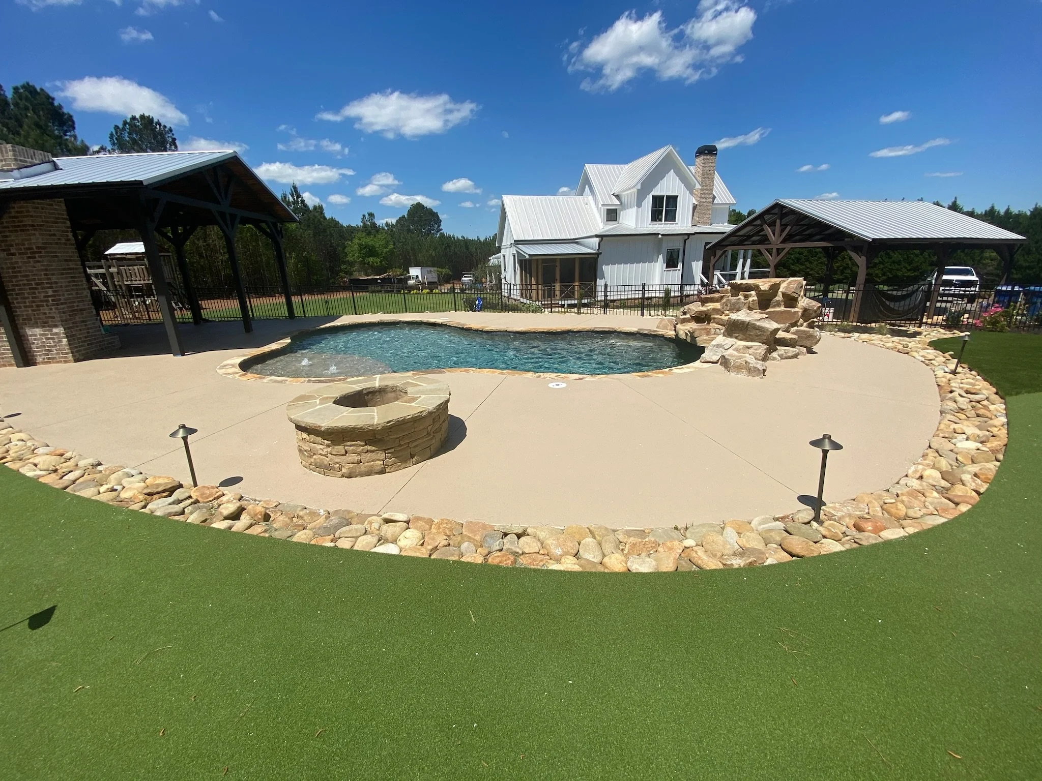 A backyard pool area with a concrete deck, surrounded by a border of small rocks, with a grassy area and a house in the background under a blue sky with a few clouds.