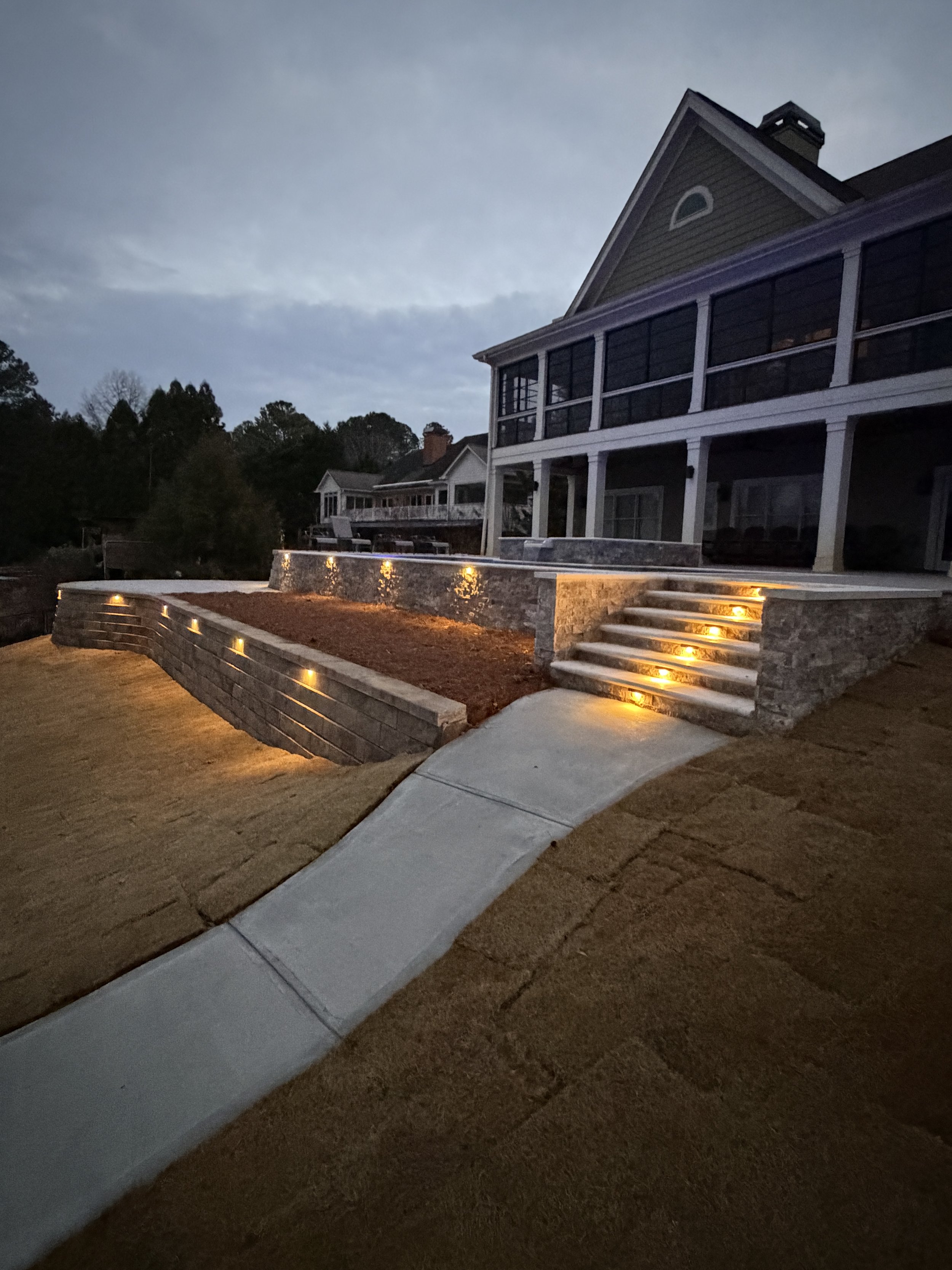 Residential house with a multi-level outdoor patio illuminated by built-in lights, a staircase leading up to the patio, and a winding concrete walkway, during dusk or early evening.