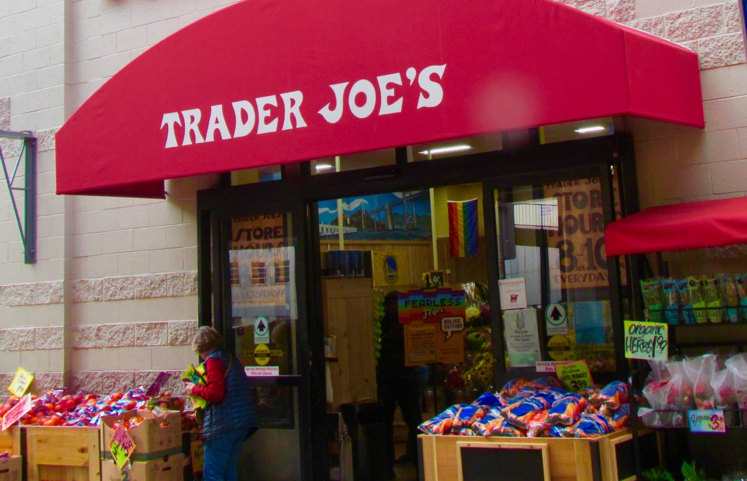 Front entrance of Trader Joe's store with red awning and signage, fruits and vegetables displayed outside.