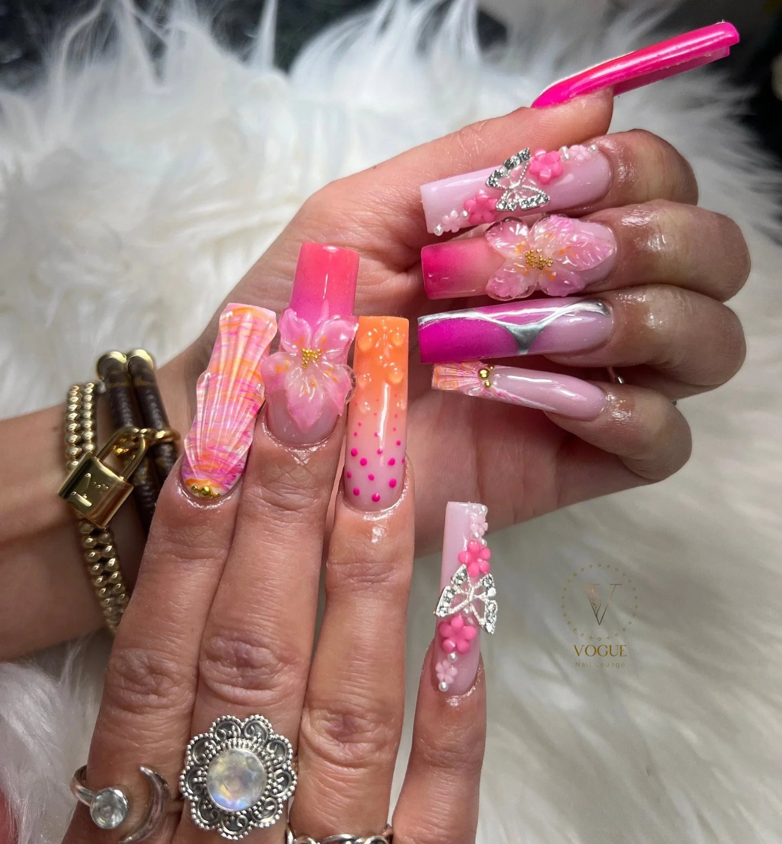 Close-up of hands with elaborate pink, orange, and purple nail art featuring floral designs, rhinestones, and 3D embellishments.