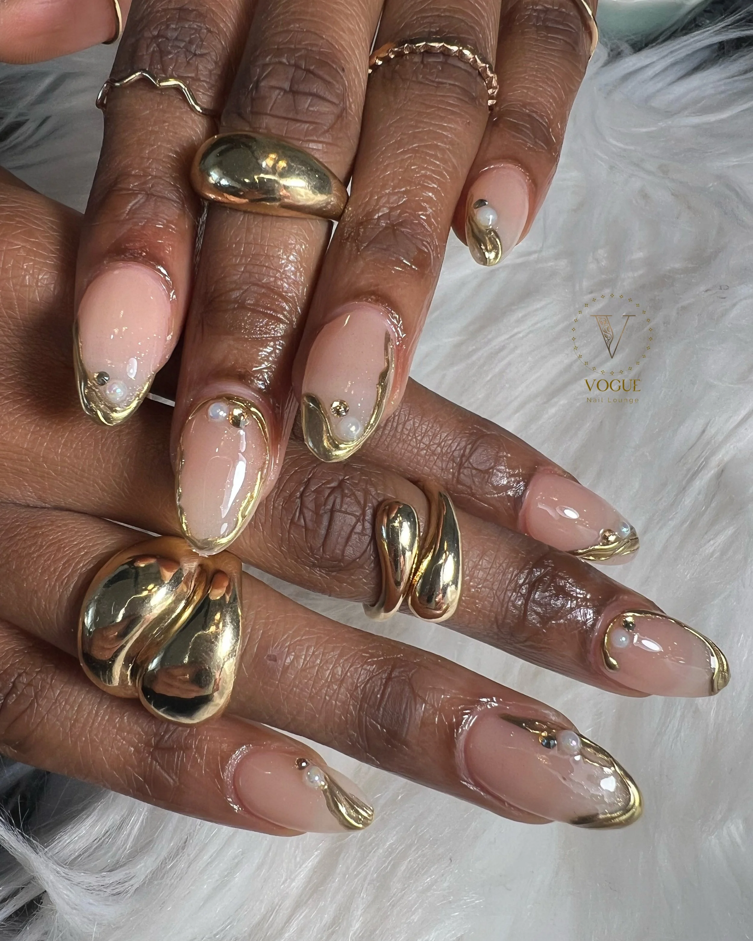 Close-up of well-manicured hands with nude nails accented with gold tips and pearl embellishments, adorned with large gold statement rings. 