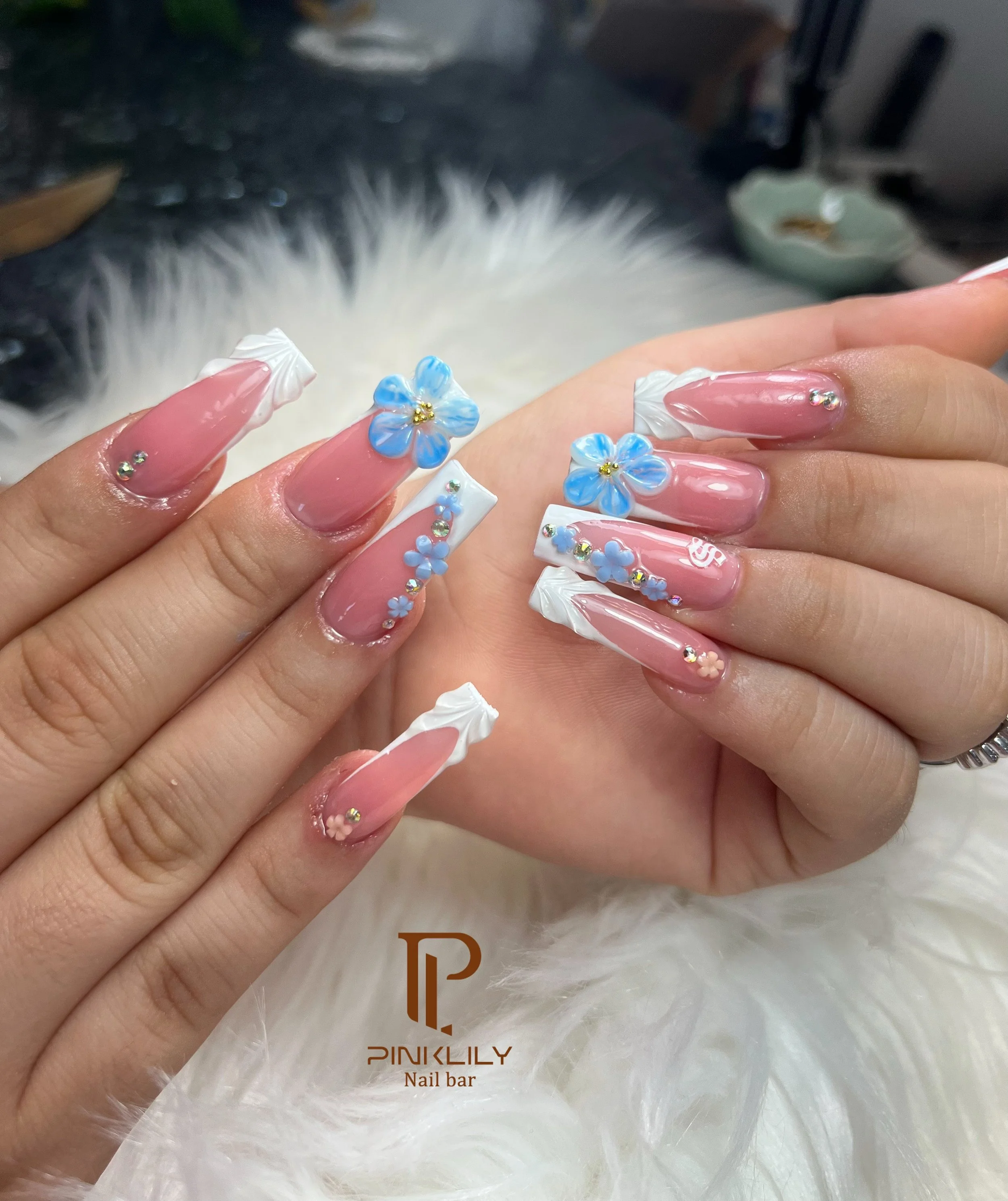 Close-up of decorated acrylic nails with pink, white, and blue accents, featuring floral designs and rhinestones.