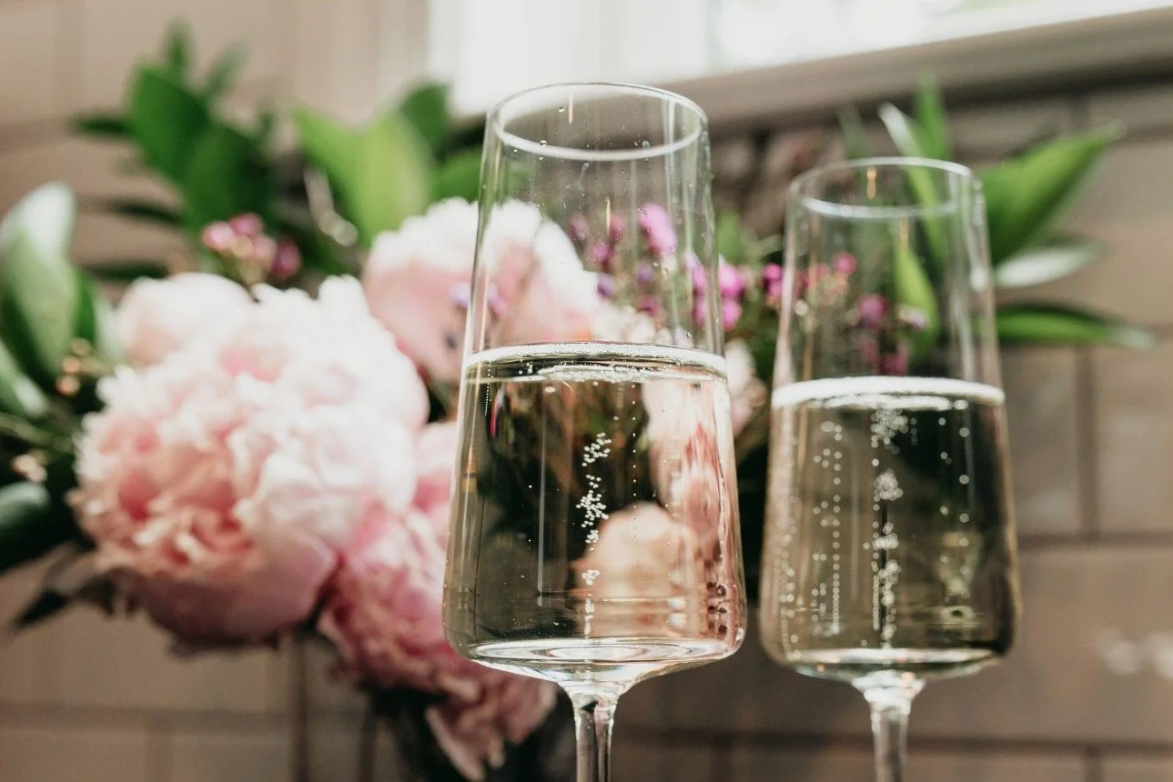 Two champagne flutes filled with sparkling water in front of floral arrangement with pink and white flowers and green leaves.
