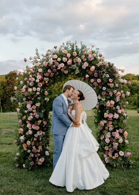Couple in wedding attire sharing a kiss under a floral arch with pink and purple roses, outdoors on a grassy field, under a cloudy sky.