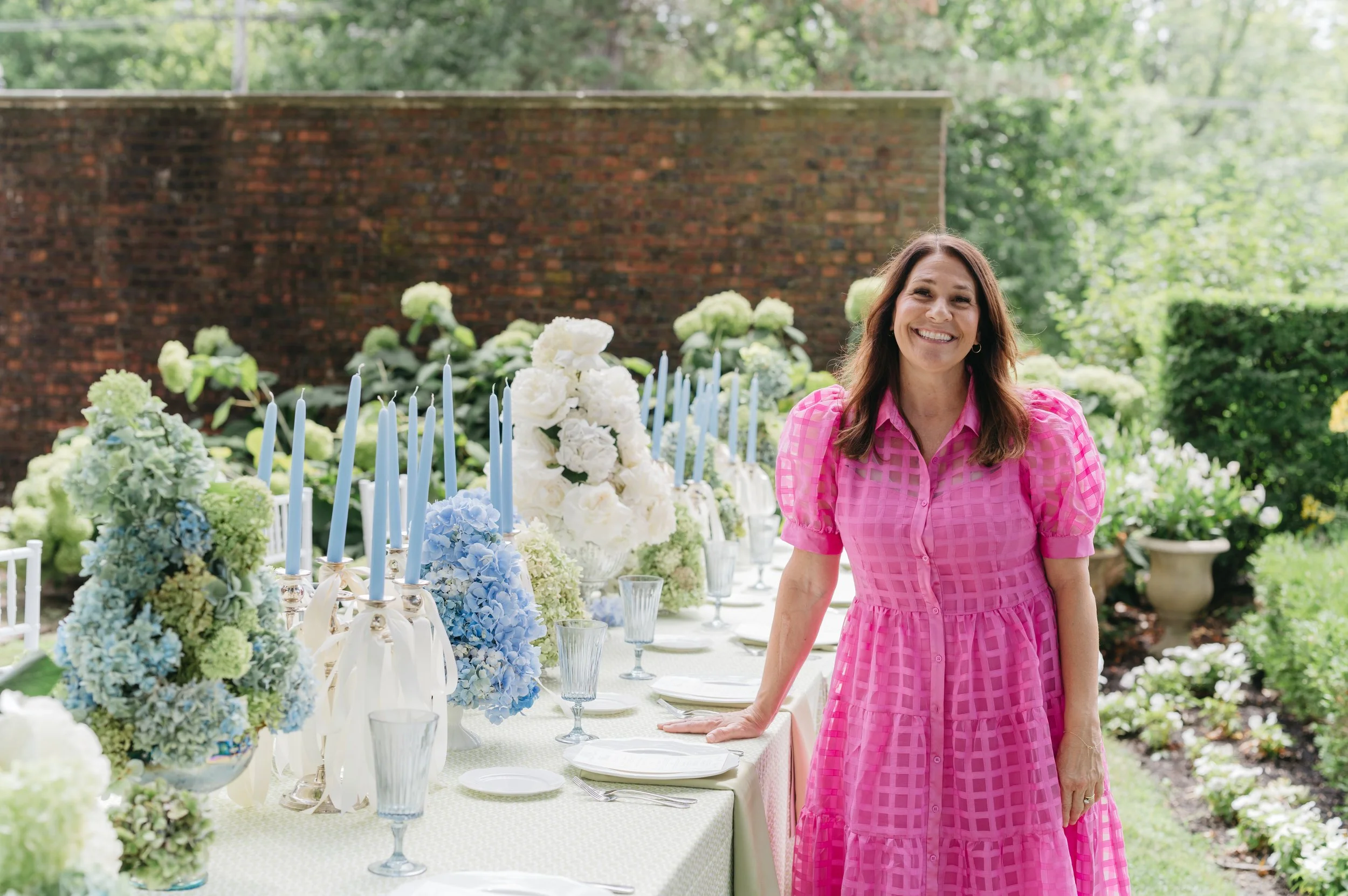 A woman in a pink dress standing outdoors beside a decorated table with blue candles, floral arrangements, and tableware in a garden setting.