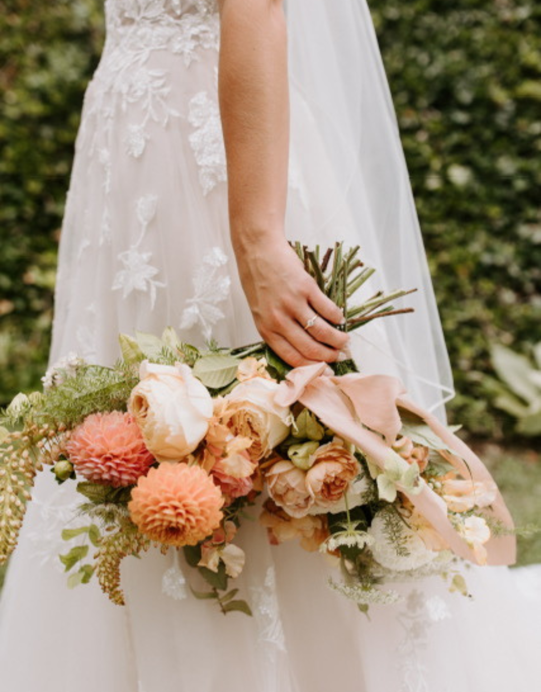 Bride holding a bouquet of blush and peach flowers, wearing a lace wedding dress, outdoors with greenery in the background.