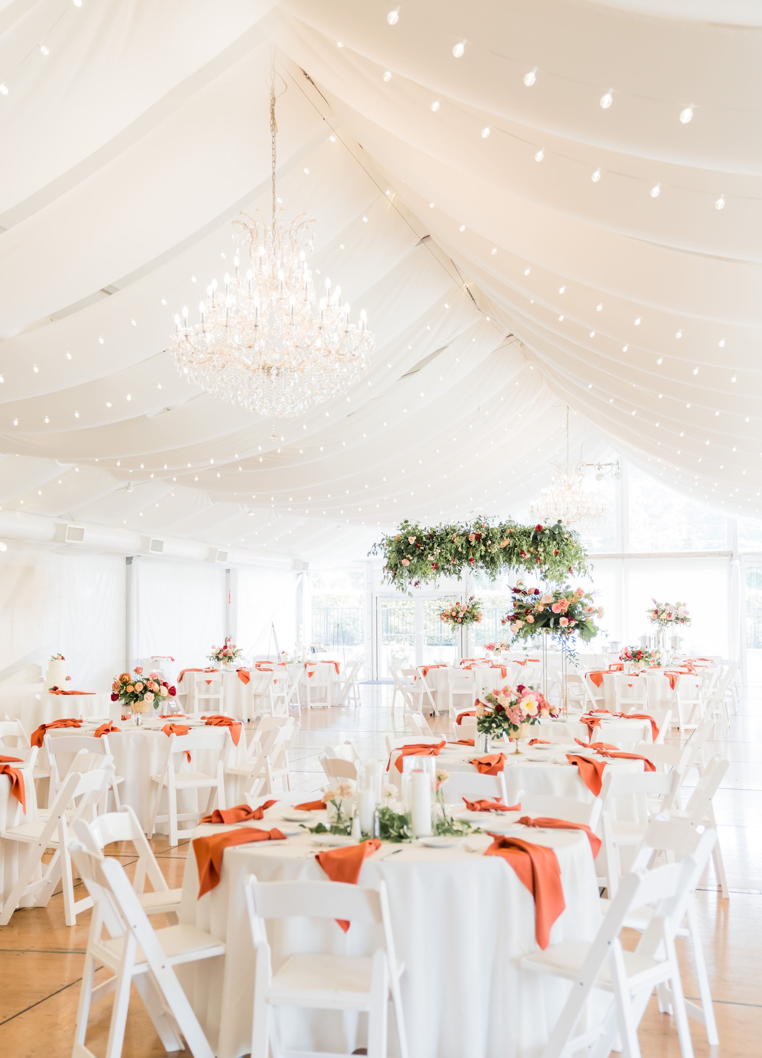 A decorated banquet hall with white tables and chairs, adorned with floral centerpieces and orange napkins, under a draped white ceiling with string lights and chandeliers.