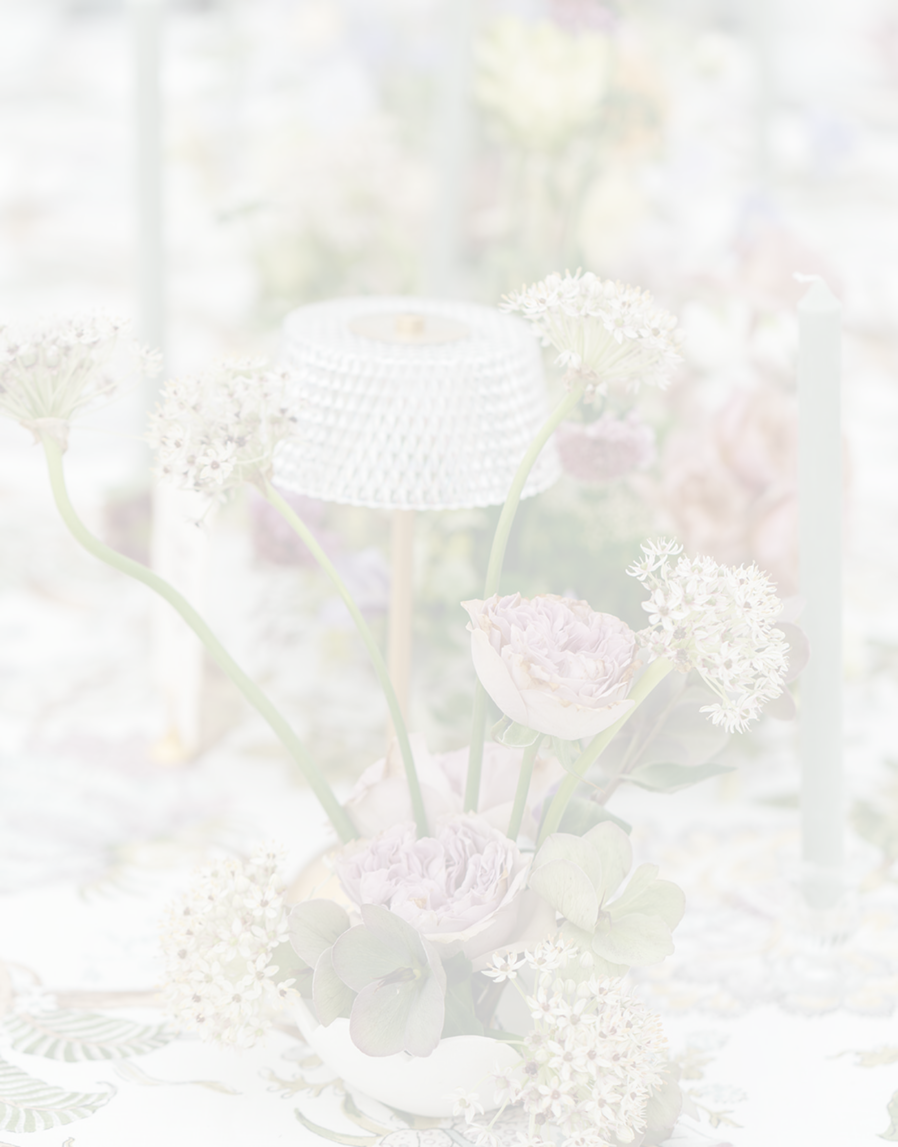 Blurred image of a floral table centerpiece with white and pale purple flowers, a white tablecloth, and a small woven lamp in the background.