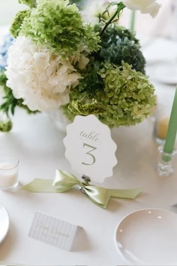 A table centerpiece with white and green hydrangeas in a vase, a table number card with 'table 3', a green candle, and place settings, all decorated with a light green ribbon.