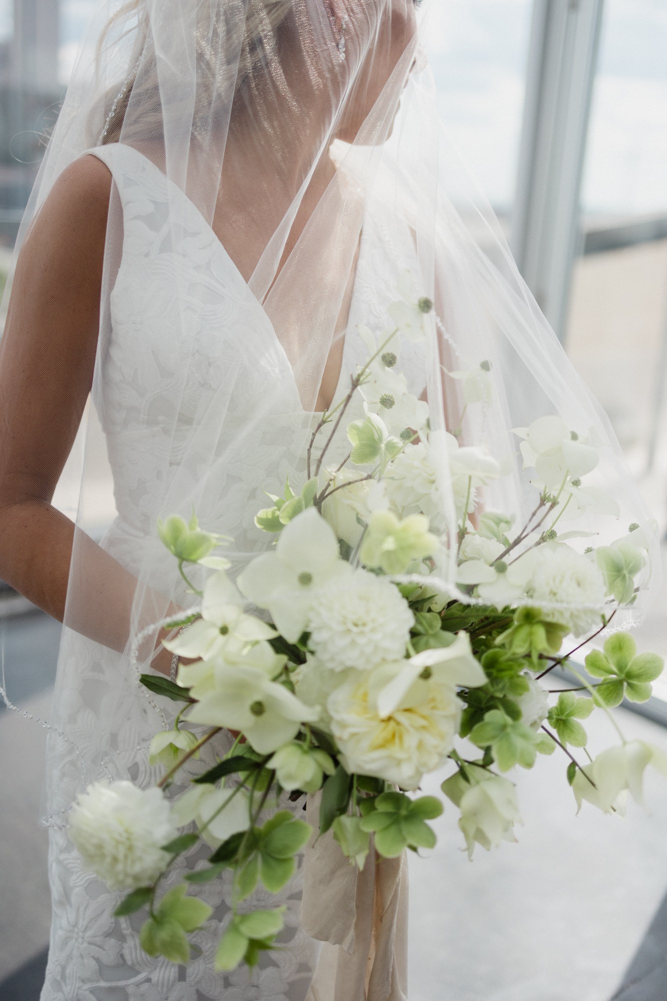 A bride in a white wedding dress holding a bouquet of white flowers, standing near a window with a sheer veil covering her face.