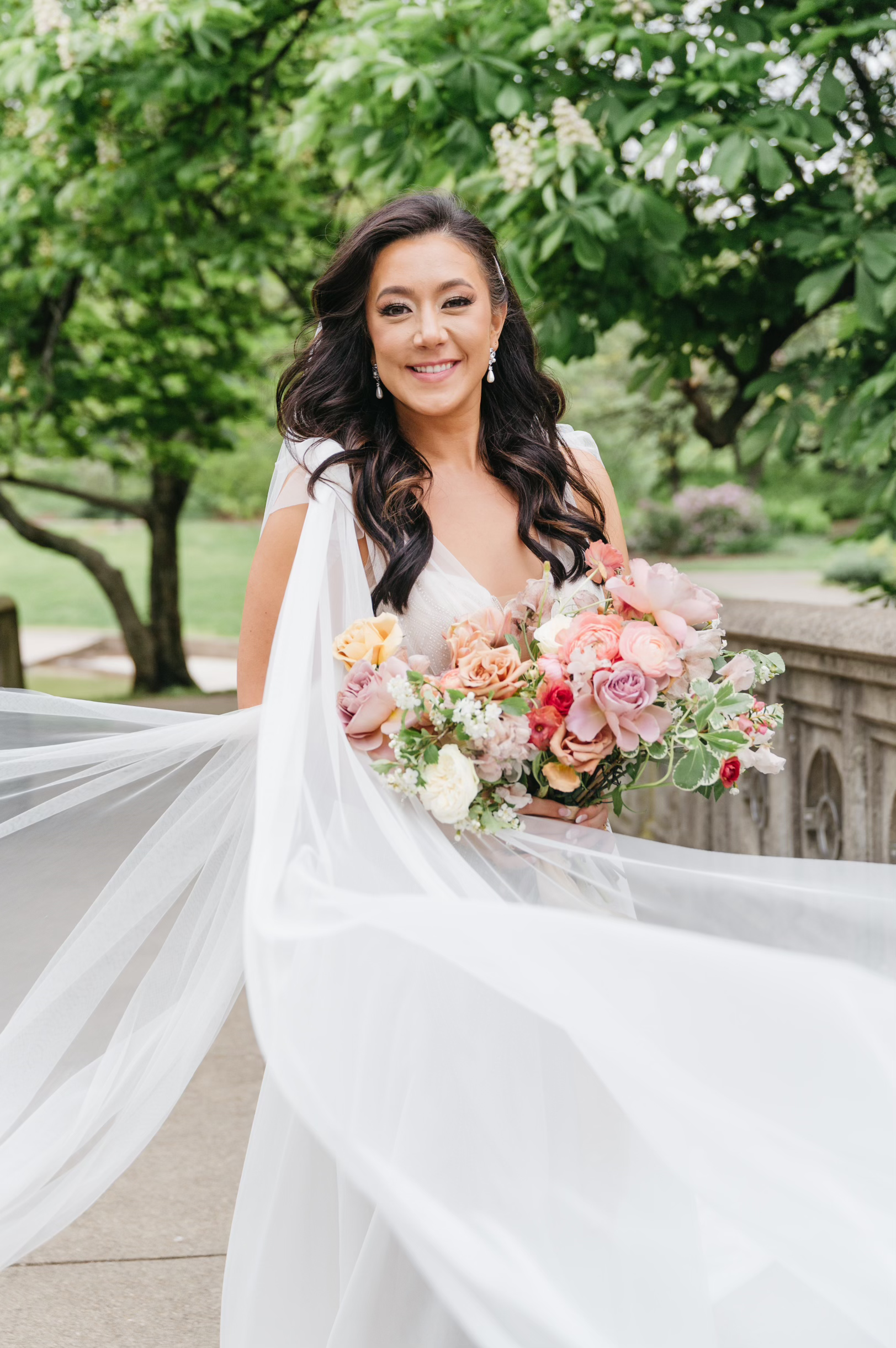 Bride holding a bouquet of pink, peach, and white flowers outdoors, wearing a white wedding dress and smiling.