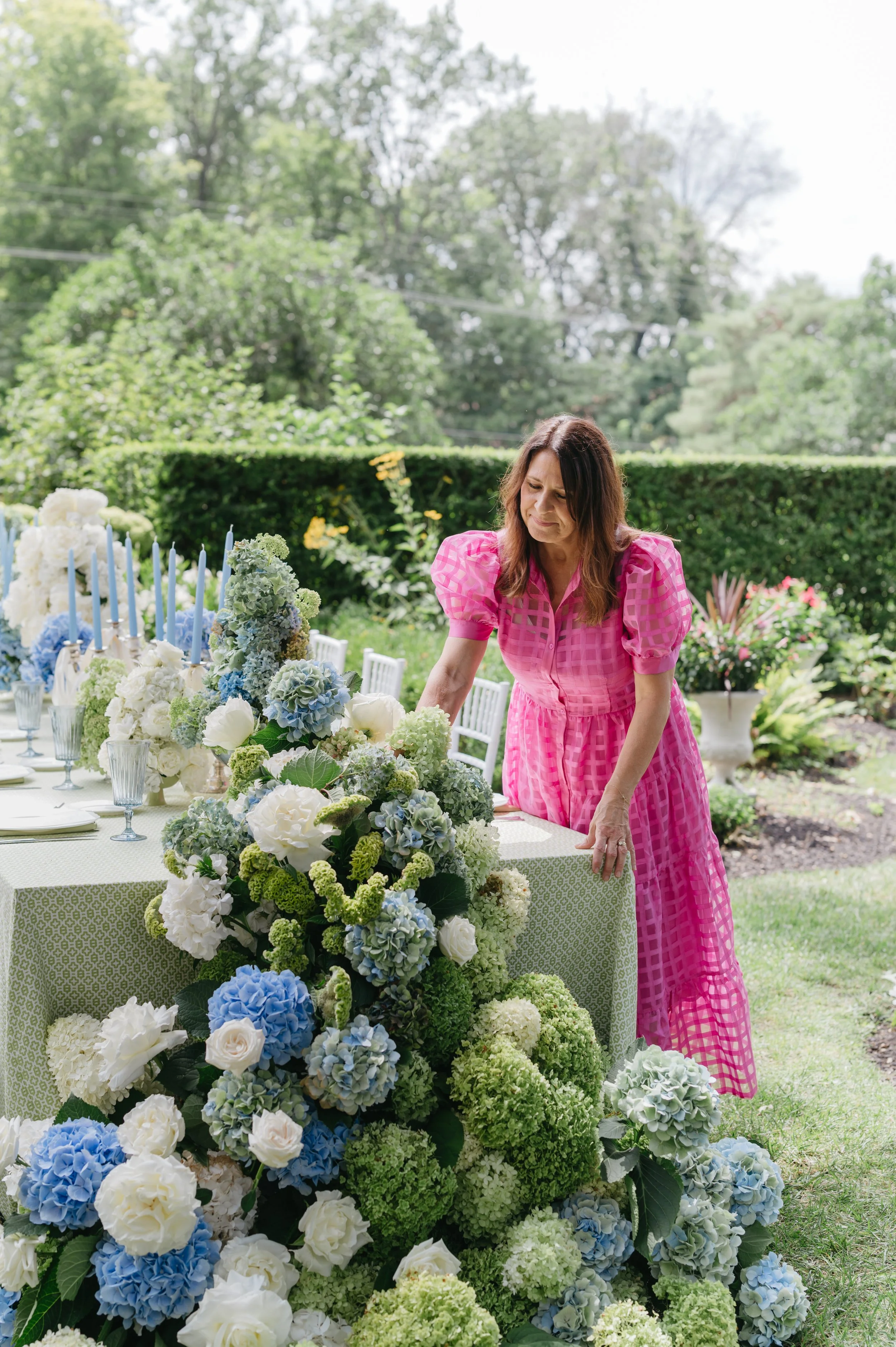 A woman in a pink dress arranging a floral centerpiece on an outdoor table set for a gathering, with outdoor greenery and trees in the background.