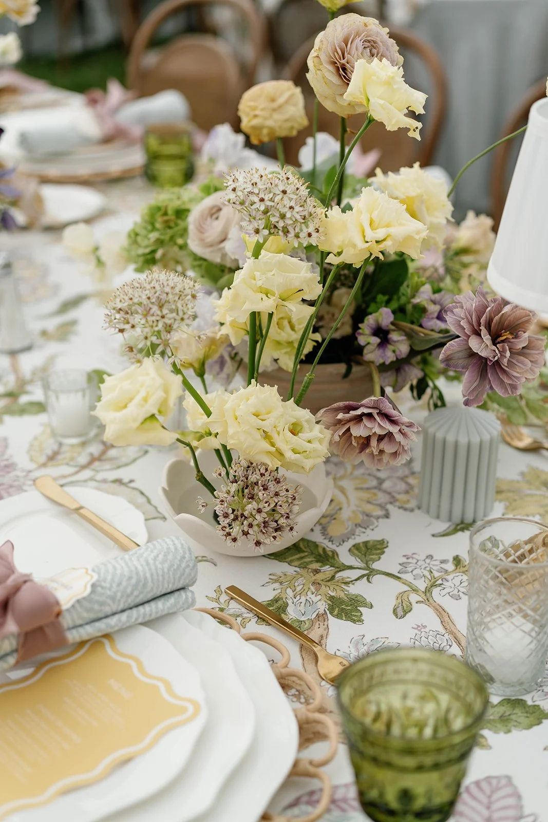A table setting with a floral centerpiece of cream, purple, and beige flowers in a white vase, gold cutlery, pastel-colored napkins, and textured glassware on a floral tablecloth.