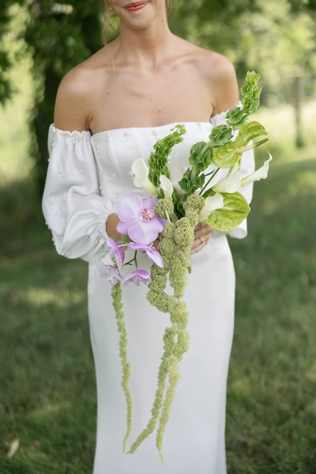 Woman in white off-the-shoulder dress holding a cascading bouquet of pink, white, and green flowers outdoors with a blurred green background.