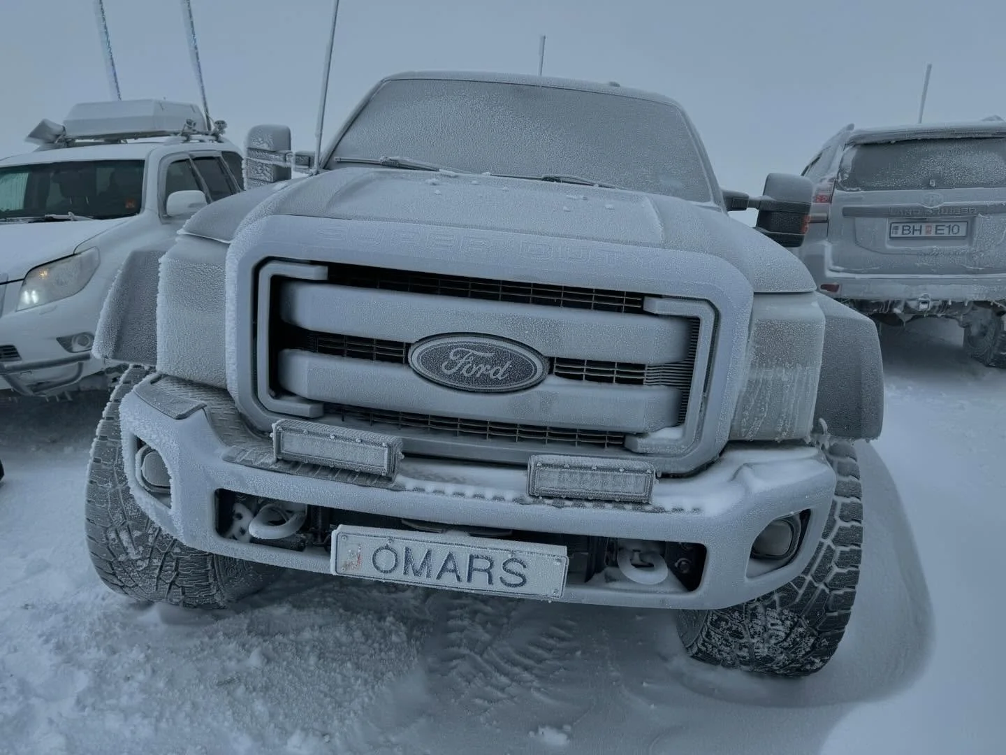 A Ford truck covered in ice and snow parked in a snowy parking lot surrounded by other vehicles, with a frosted license plate reading 'OMARS'.