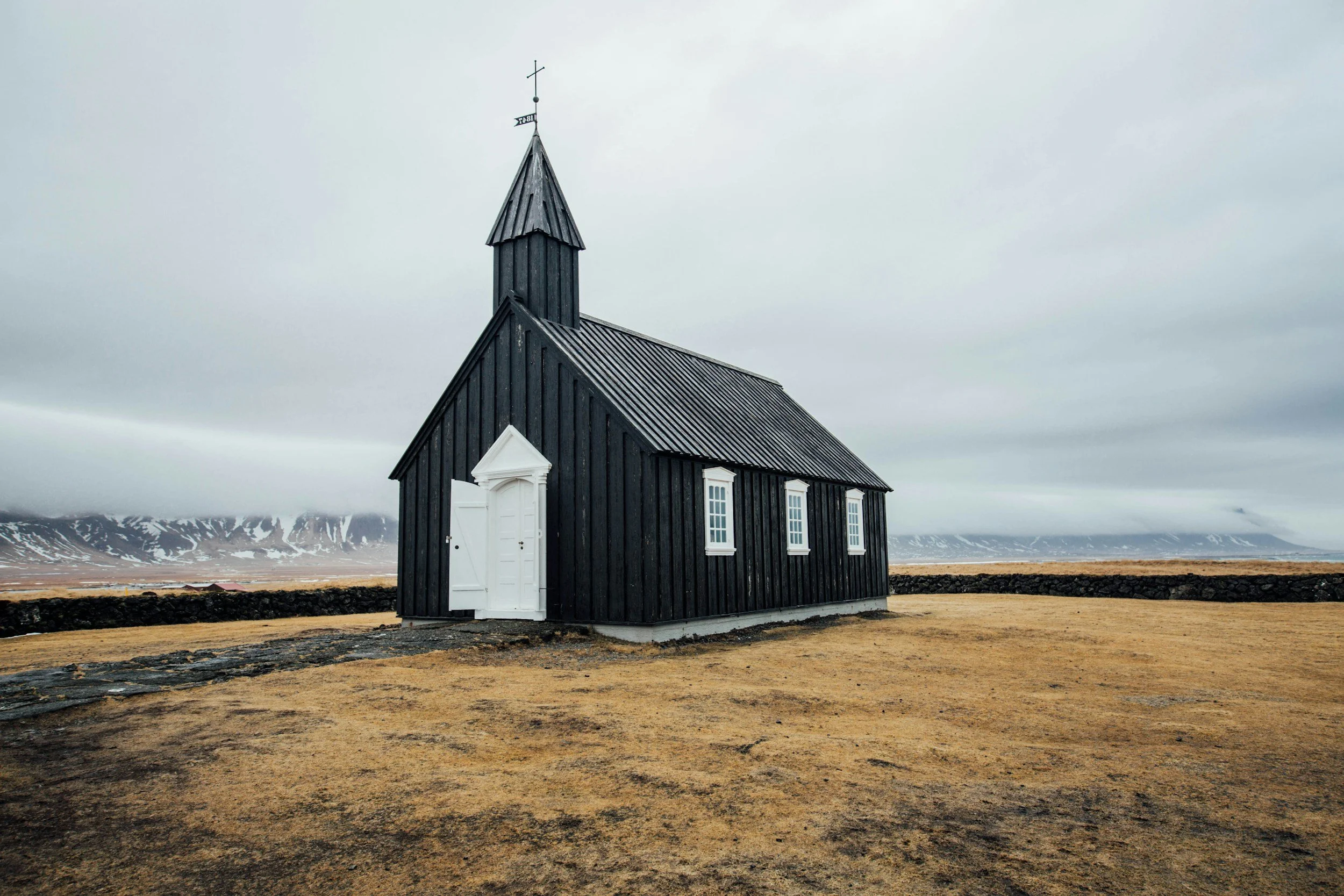 A small black wooden church with white window frames and a white door, situated on a grassy field with a stone wall in the background, under a cloudy sky.