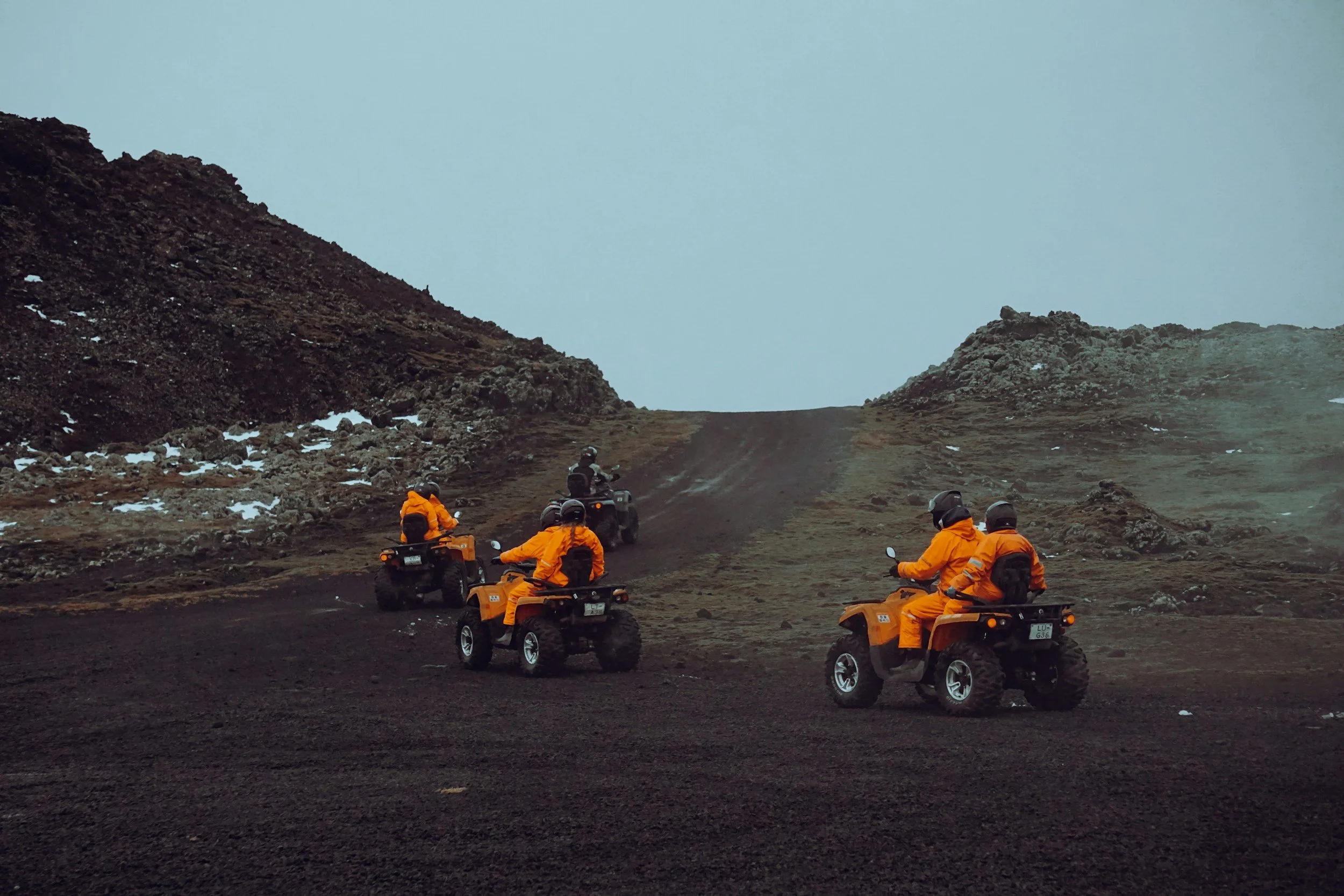 Group of five people riding all-terrain vehicles up a rugged, rocky dirt trail on a cloudy day, surrounded by mountainous terrain.