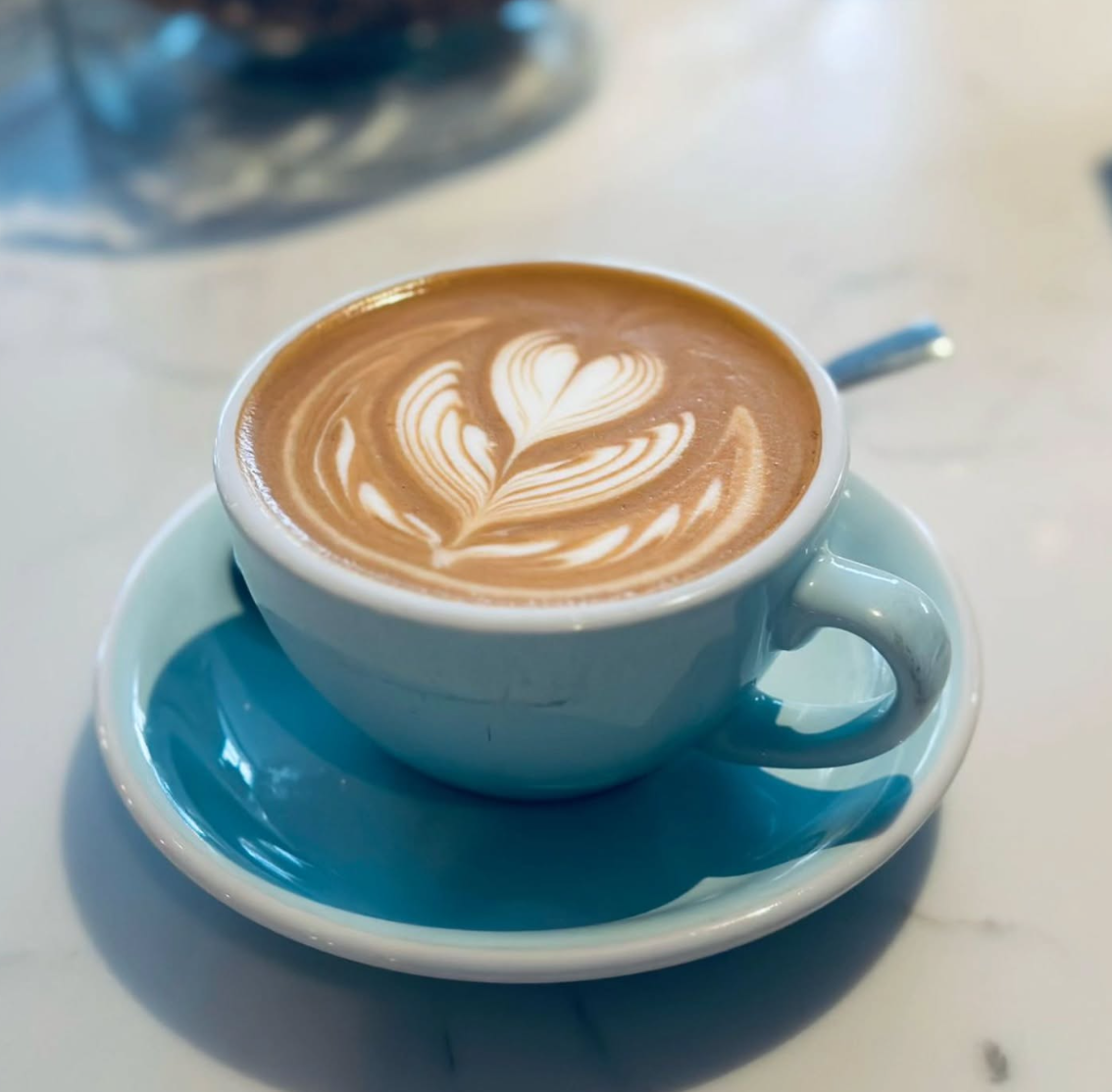 A white cup of latte with heart latte art on top, sitting on a light blue saucer on a white table.