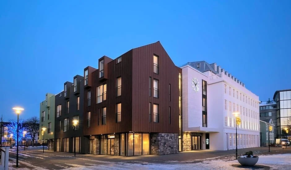 Modern multi-story buildings on a city street at dusk, with illuminated windows and street lamps.