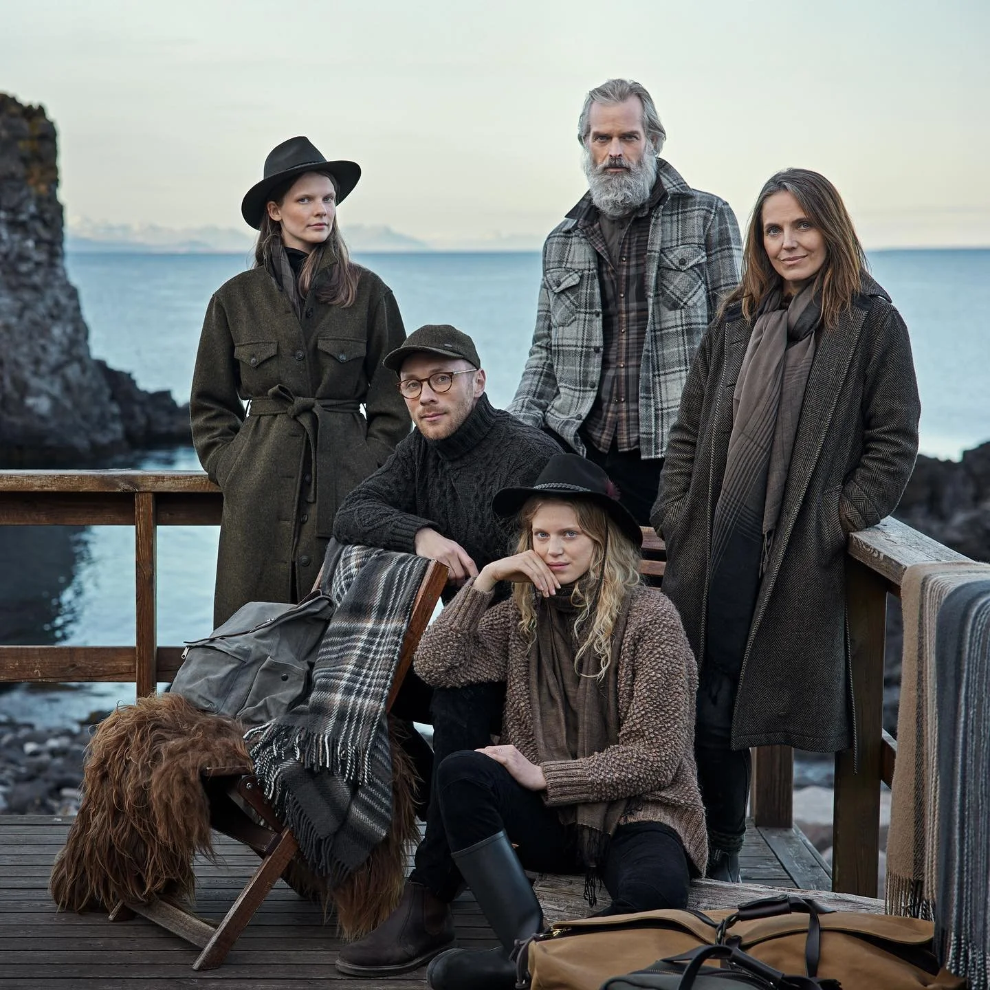 Group of six people and a dog on a wooden deck by the sea, dressed in fall or winter clothing.