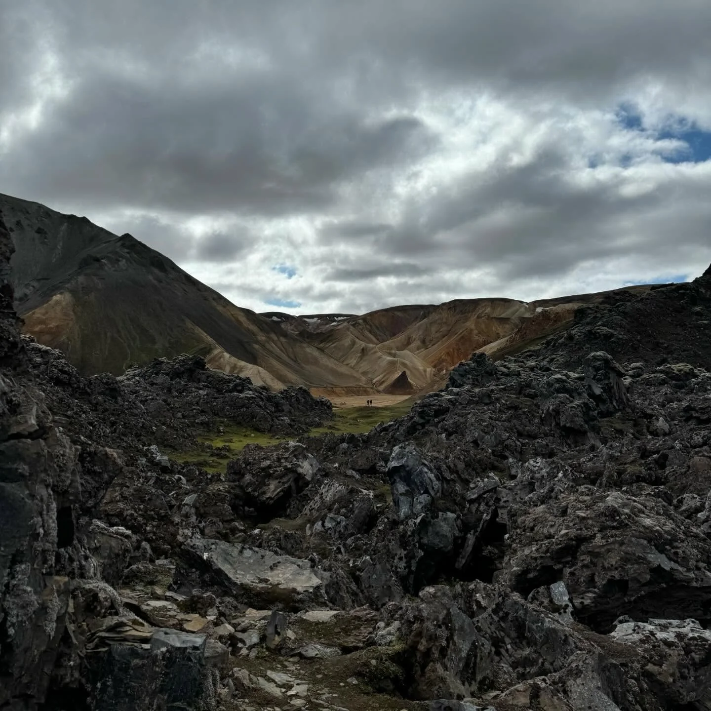 A rocky volcanic landscape with dark lava rocks in the foreground, multicolored mountains distant, and a cloudy sky above.