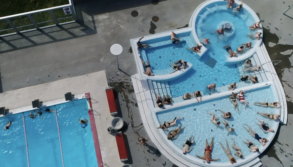 Aerial view of a swimming pool area with multiple pools. Several people are swimming and lounging in the pools on a sunny day.