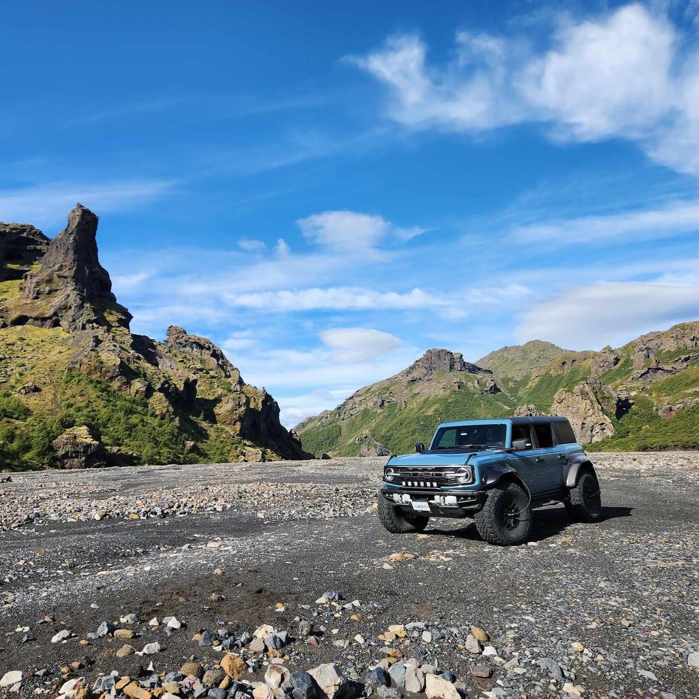 A vintage blue off-road vehicle parked on rocky terrain with green mountains and a blue sky with clouds in the background.
