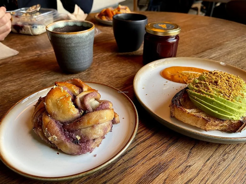 Fried apple fritter on a white plate, sliced avocado with crushed topping on toast with sauce, coffee, and jam jars on a wooden table.