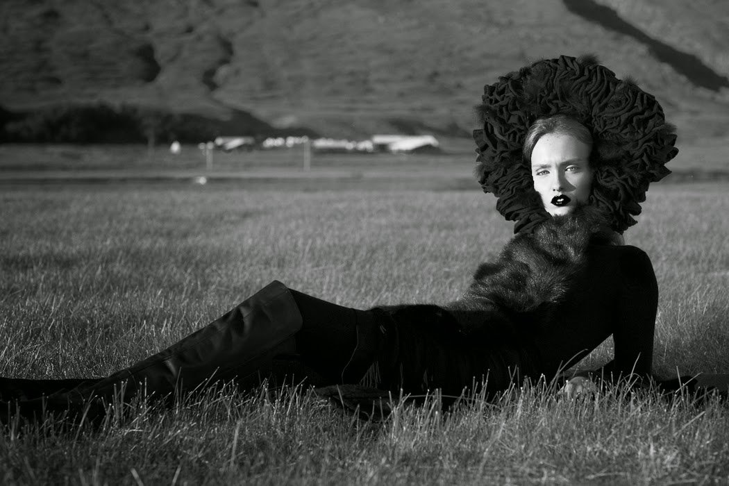 A woman lying on the grass in a field, wearing a large ruffled hat and dark clothing, with mountains in the background.