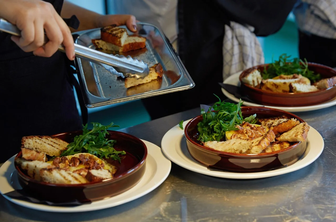 People serving and preparing plates of grilled sandwiches with greens at a restaurant table.