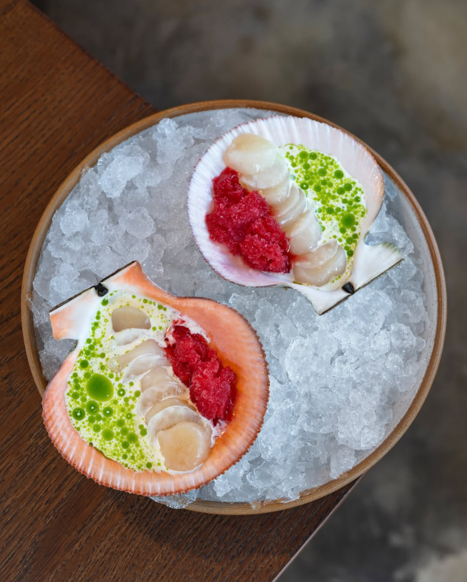 Two seashells filled with scallops, red sauce, green sauce, and oil on ice in a wooden bowl.