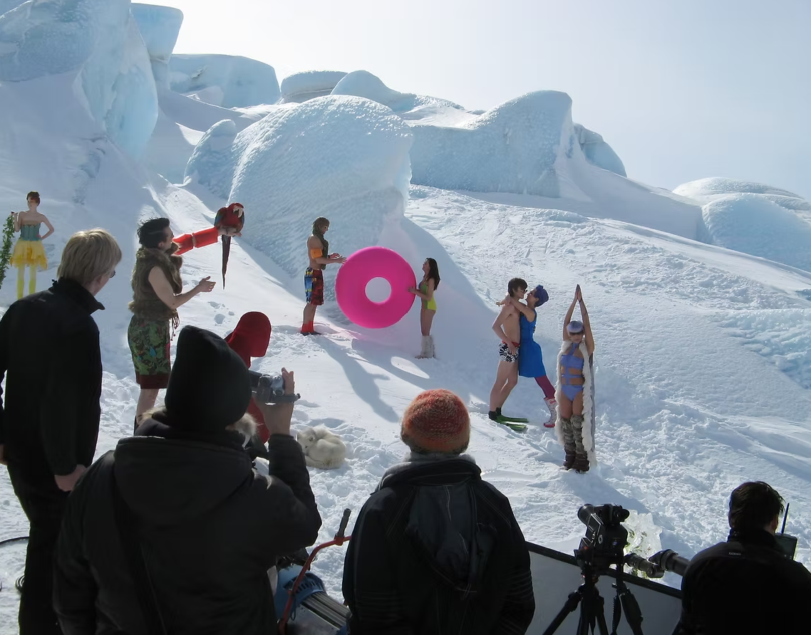People dressed in swimsuits and summer clothes on a snowy landscape with glaciers, some holding inflatable pool floats and skiers, while others are observing and taking pictures.