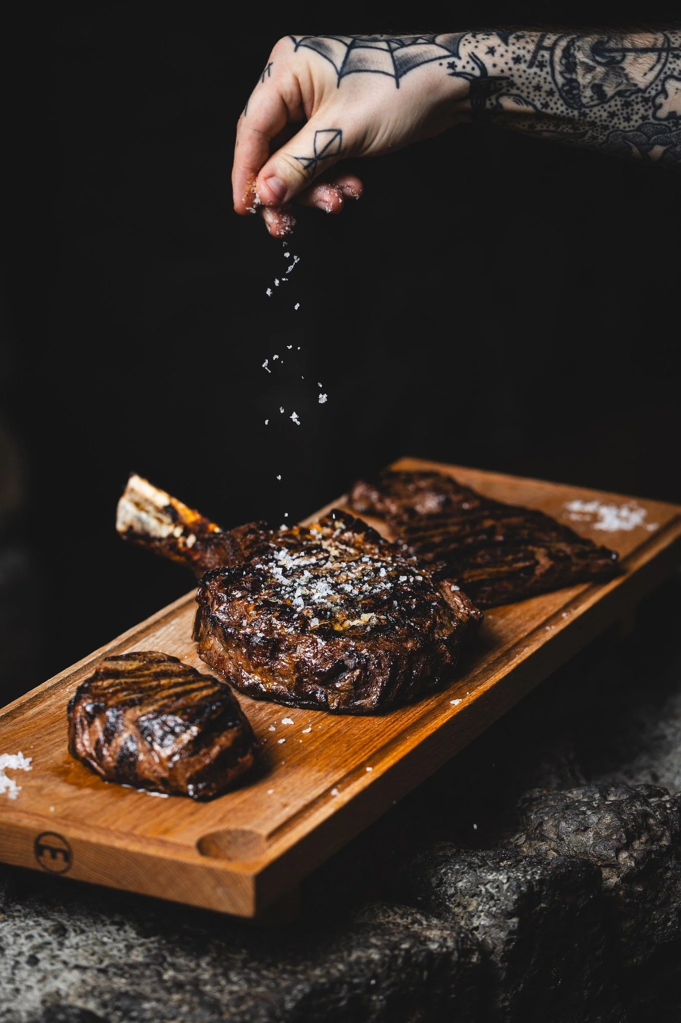 A tattooed hand sprinkling salt on grilled steaks on a wooden serving board.