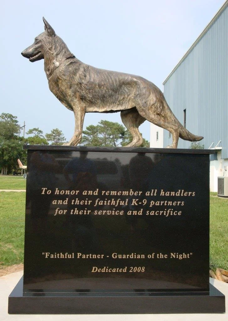 A bronze statue of a German Shepherd dog on a black pedestal with an inscription honoring K-9 police partners for their service and sacrifice, dedicated in 2008.