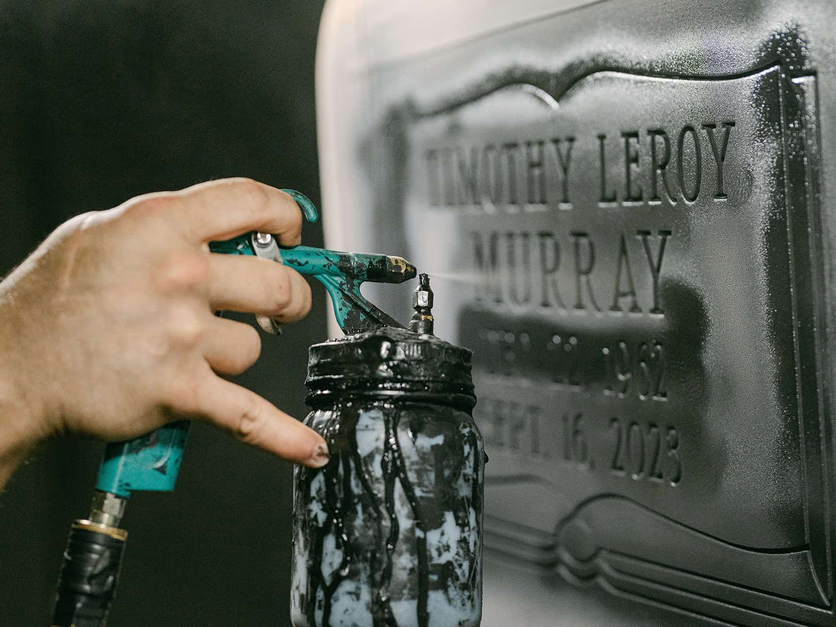 A mans hand holding a spray painting canister to an engraved headstone