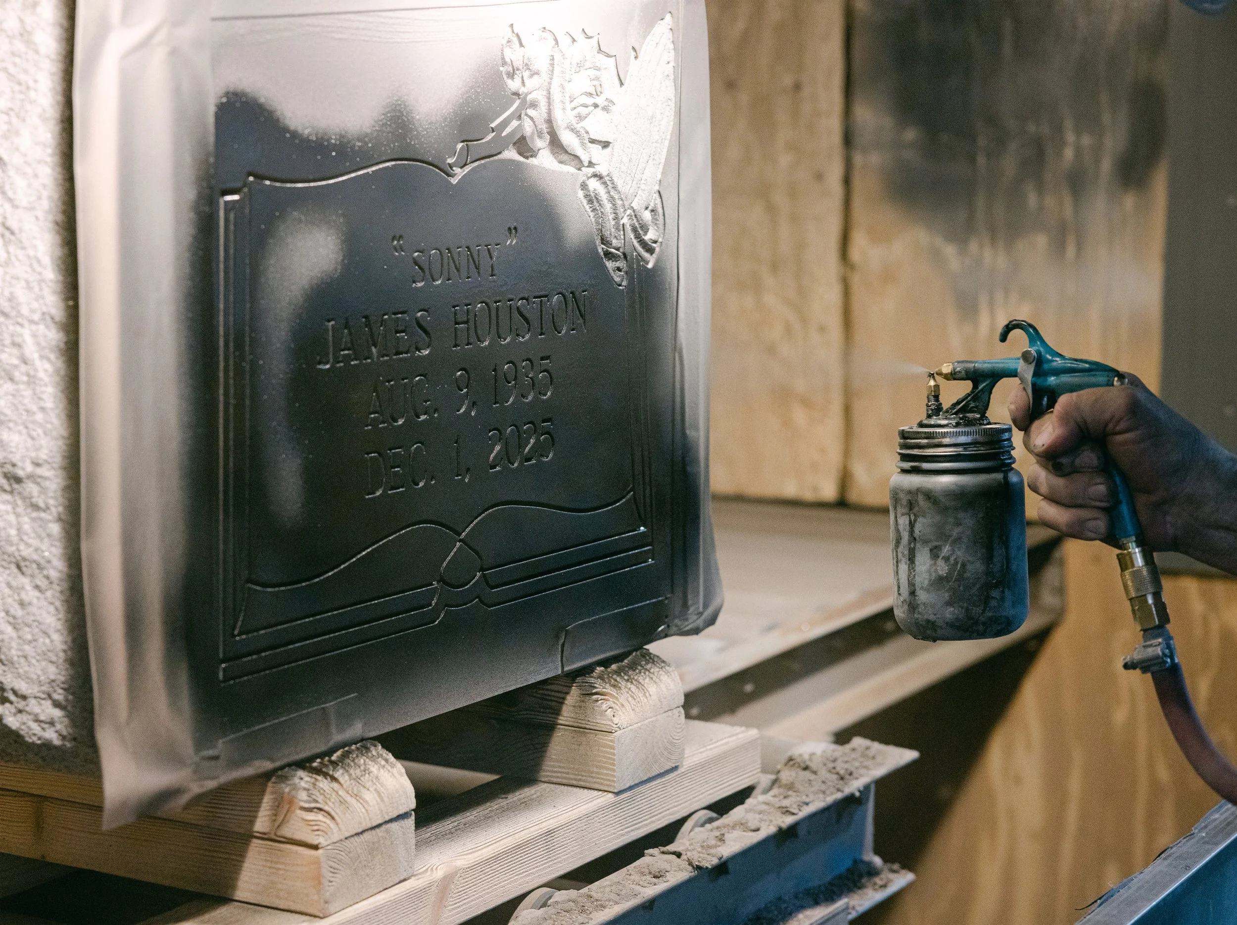 A hand spraying black paint on the text of a headstone with a stencil protecting the granite
