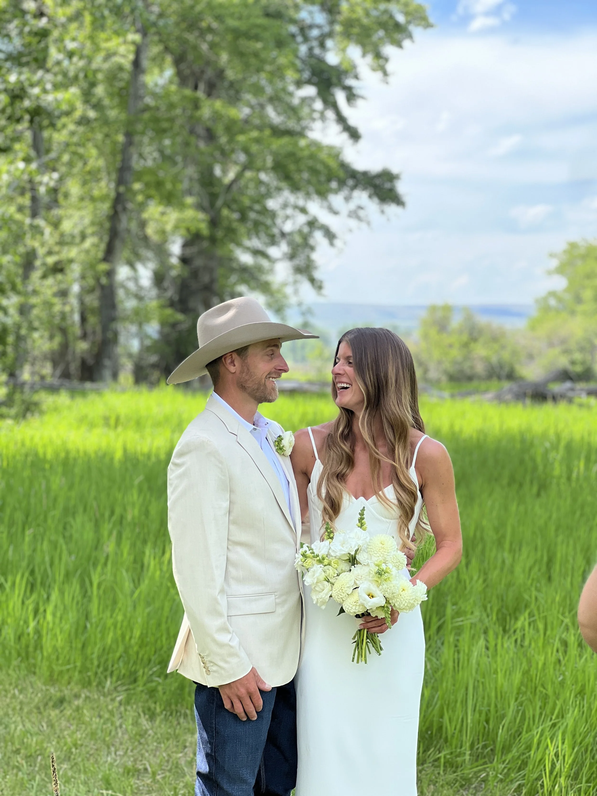 A couple is standing outdoors in a lush green field with trees and a cloudy sky, sharing a joyful moment. The man wears a light-colored suit and cowboy hat, and the woman wears a white dress and is holding a bouquet of white flowers.
