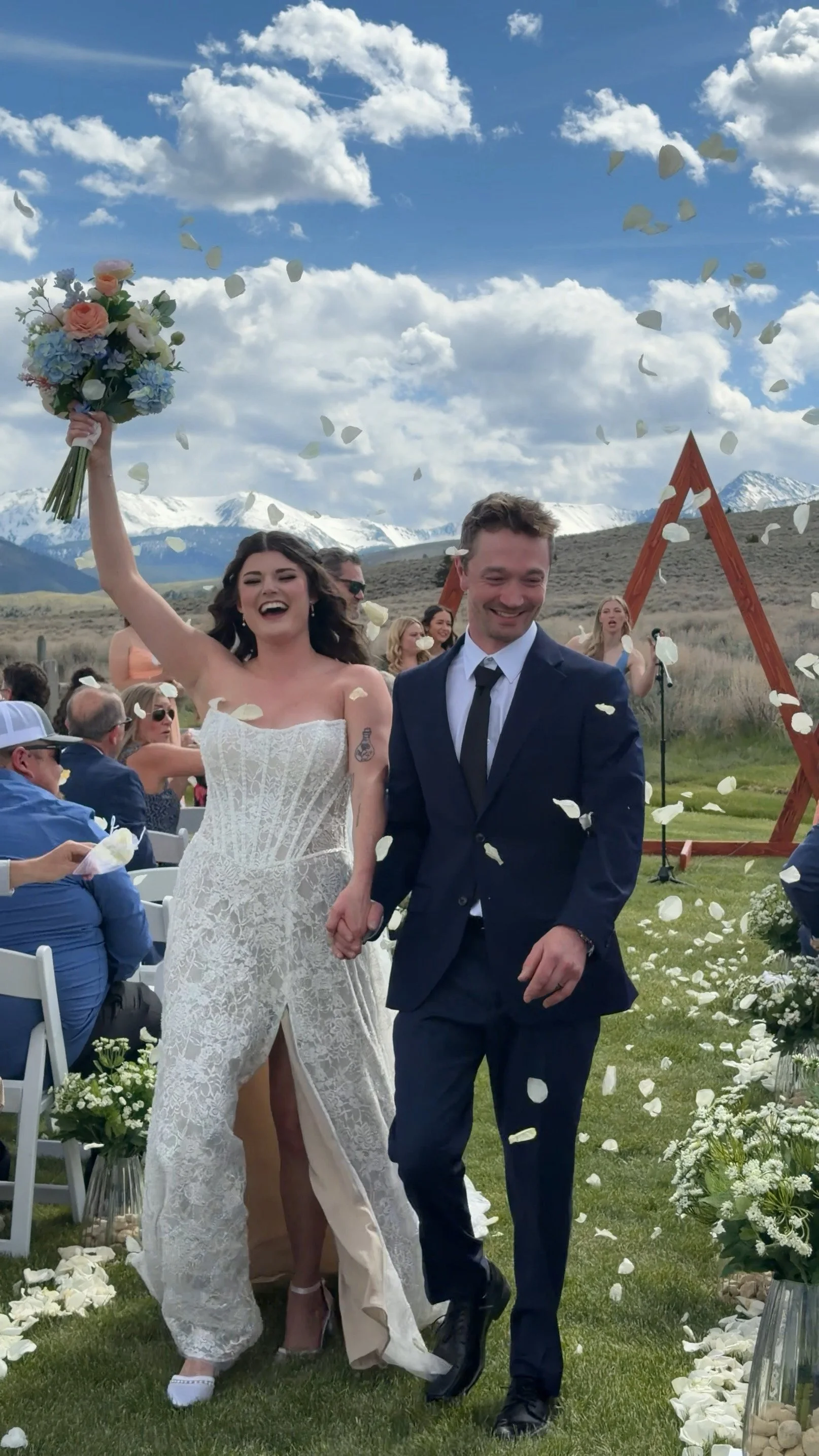 A newlywed couple walks down the aisle outdoors during their wedding ceremony, with guests in the background and mountain scenery under a partly cloudy sky, as white flower petals are thrown into the air.