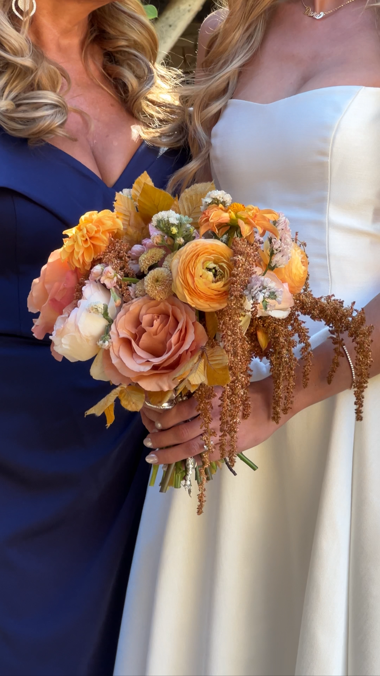 Close-up of two women at a wedding, one in a white dress holding a bouquet, the other in a dark blue dress, both with long curled blonde hair.