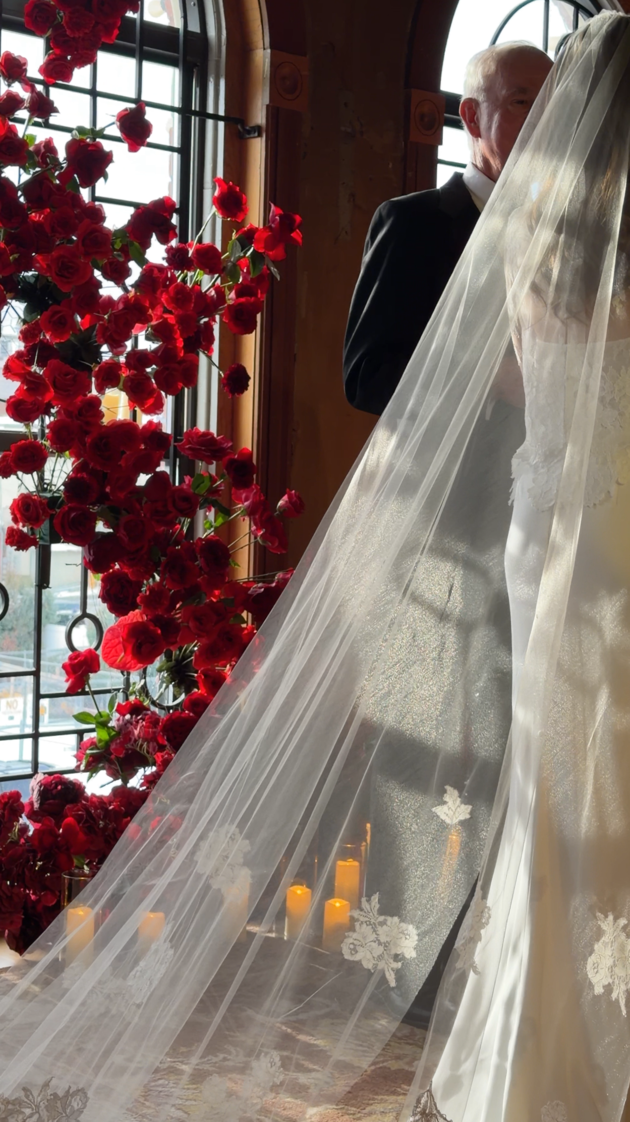 A groom and bride standing behind a sheer white veil at a wedding ceremony, with a window and red flowers in the background.