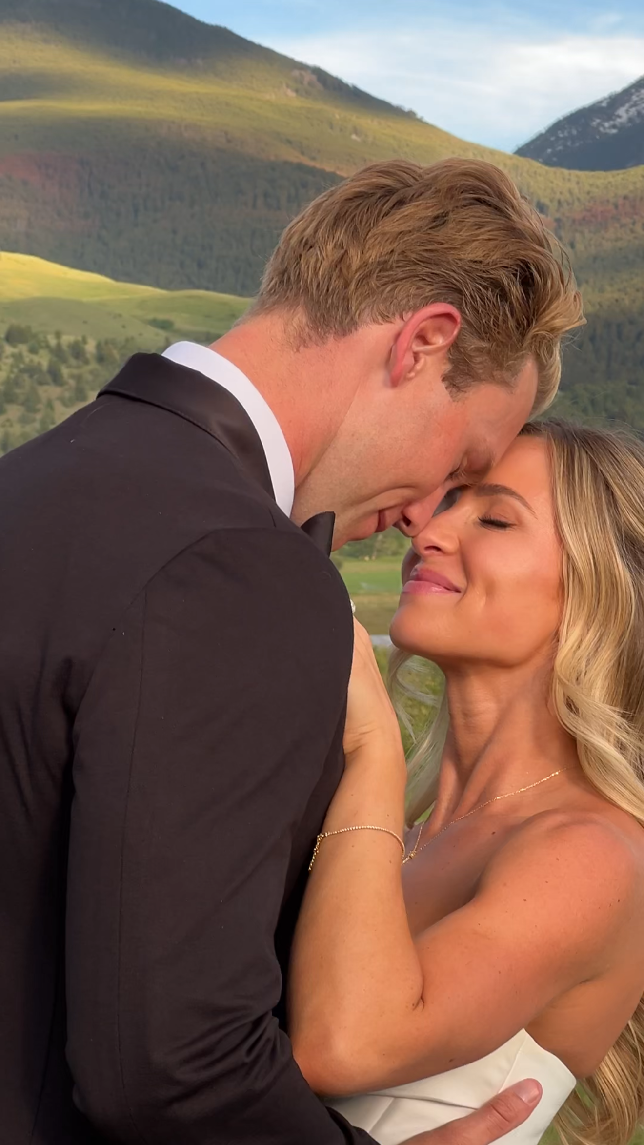 A man in a tuxedo and a woman in a white dress sharing a tender moment with their foreheads touching outdoors, with mountains and a cloudy sky in the background.
