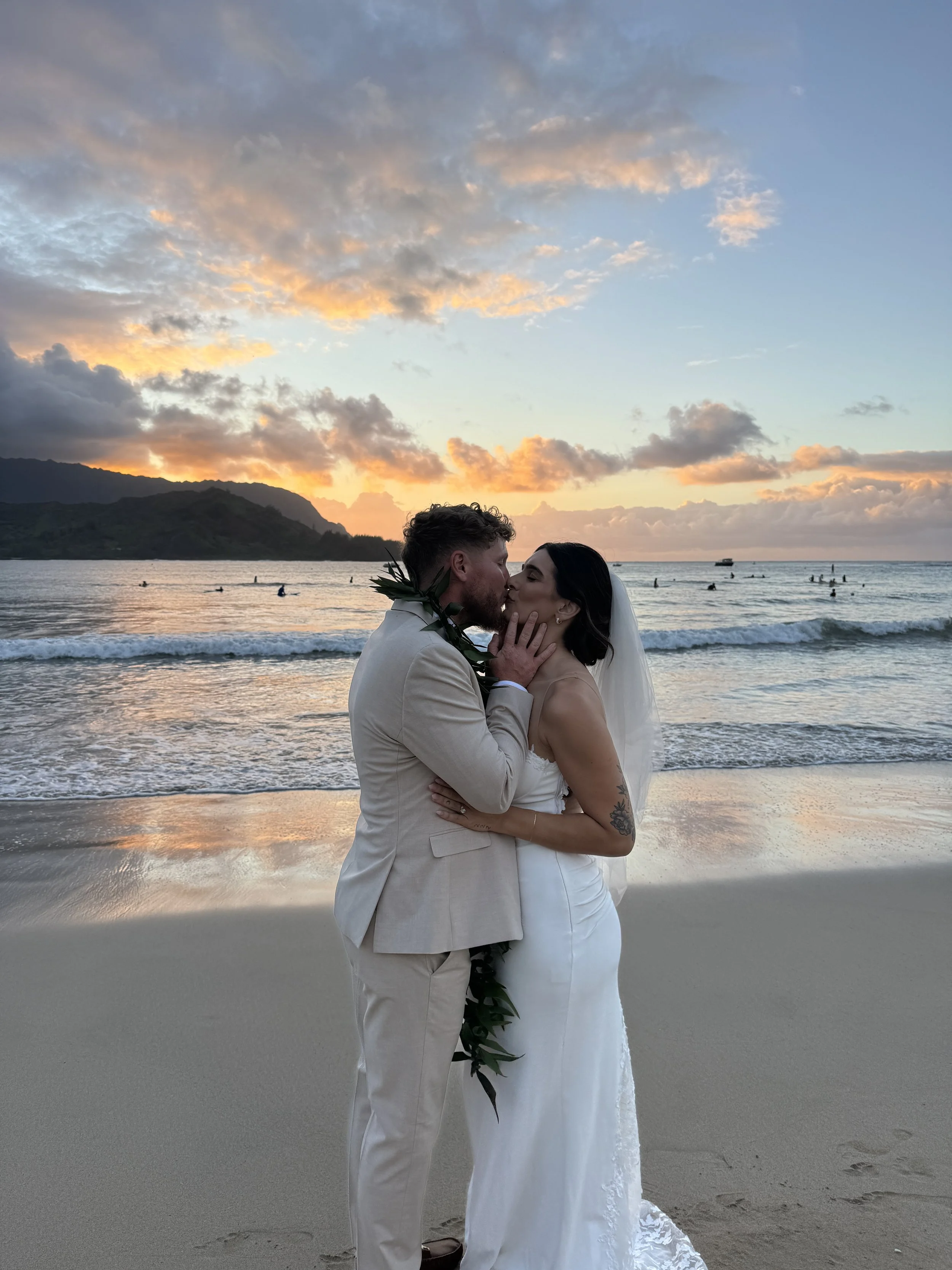 A couple in wedding attire sharing a kiss on the beach at sunset, with mountains and boats in the background.