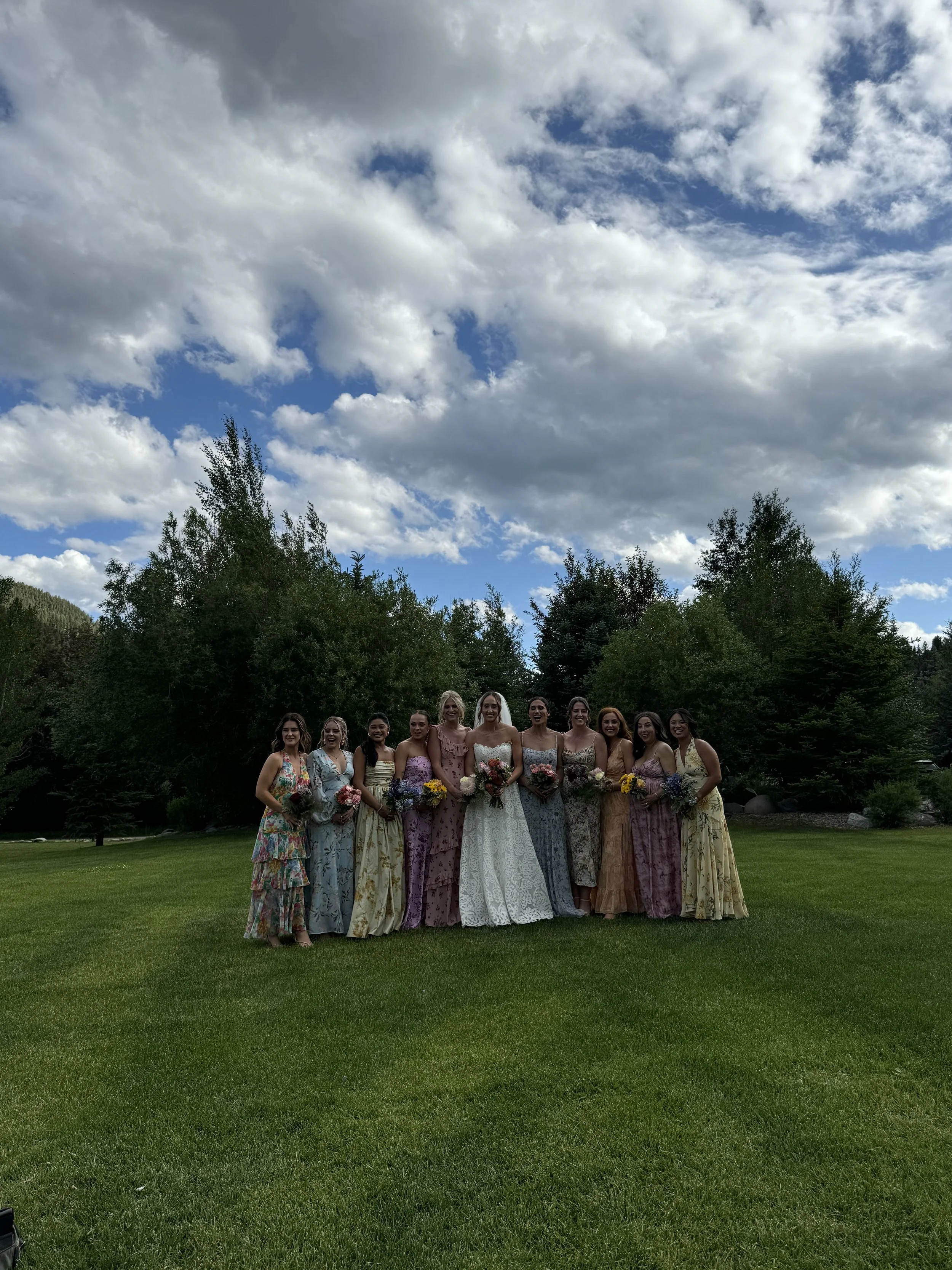 A group of women dressed in colorful, floral dresses standing on a lush green lawn outdoors with a backdrop of trees and a partly cloudy sky.