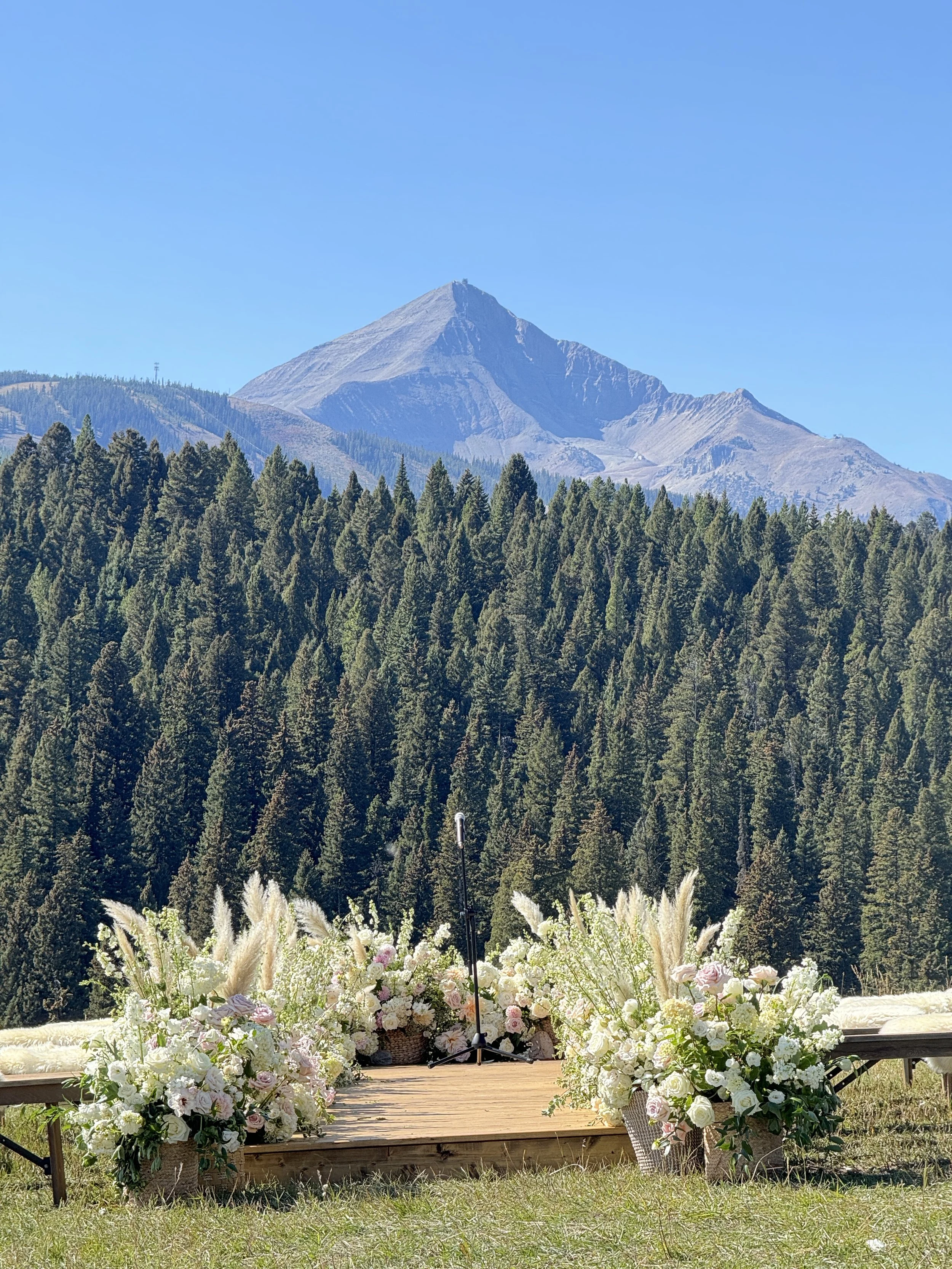 Outdoor wedding altar with white and pink flowers on a wooden platform, surrounded by tall grasses and lush green pine trees with mountain in the background under a clear blue sky.