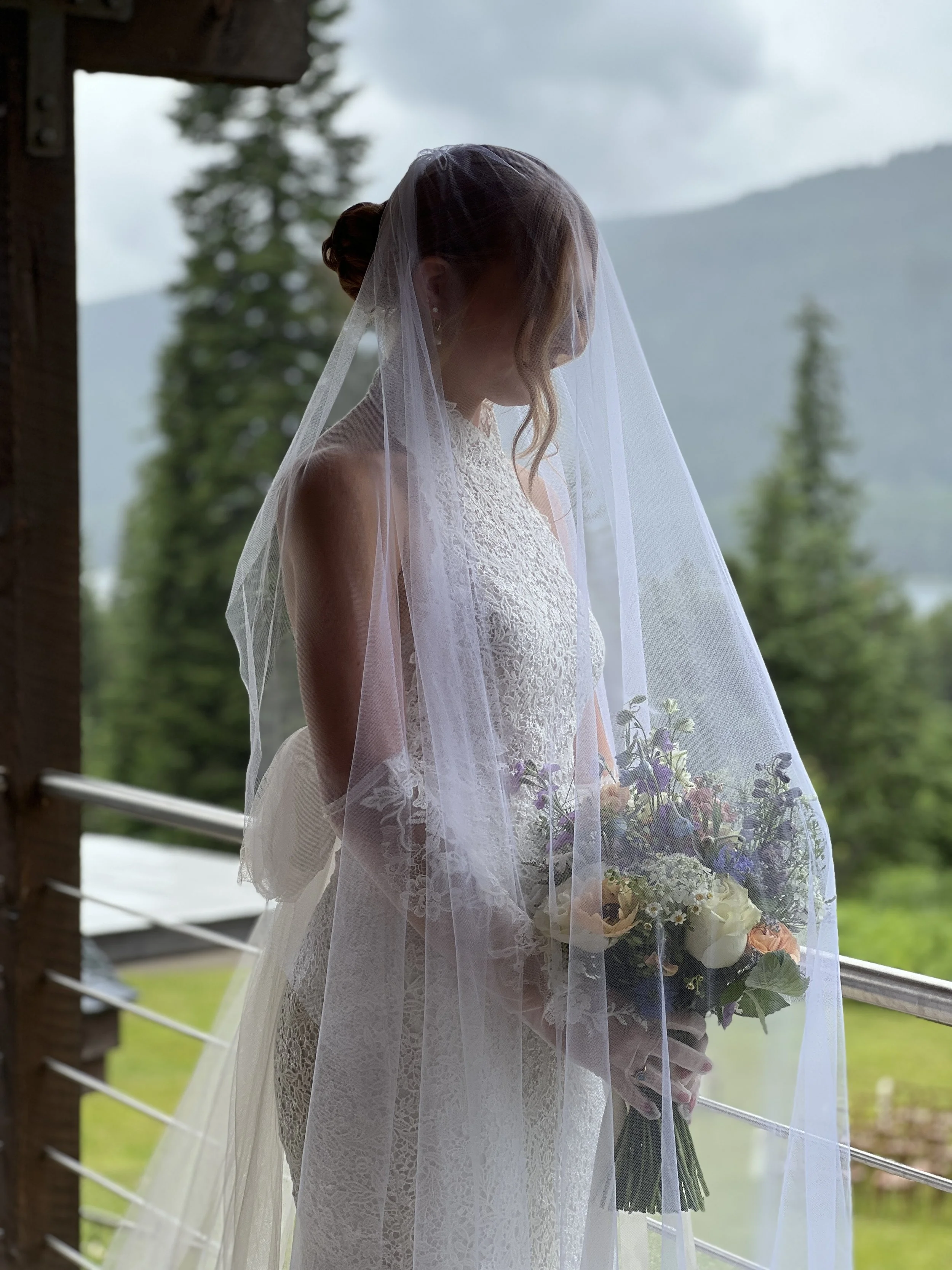A bride in a white lace wedding dress with a veil holding a bouquet of flowers, standing outdoors on a balcony with mountains and trees in the background during daytime.