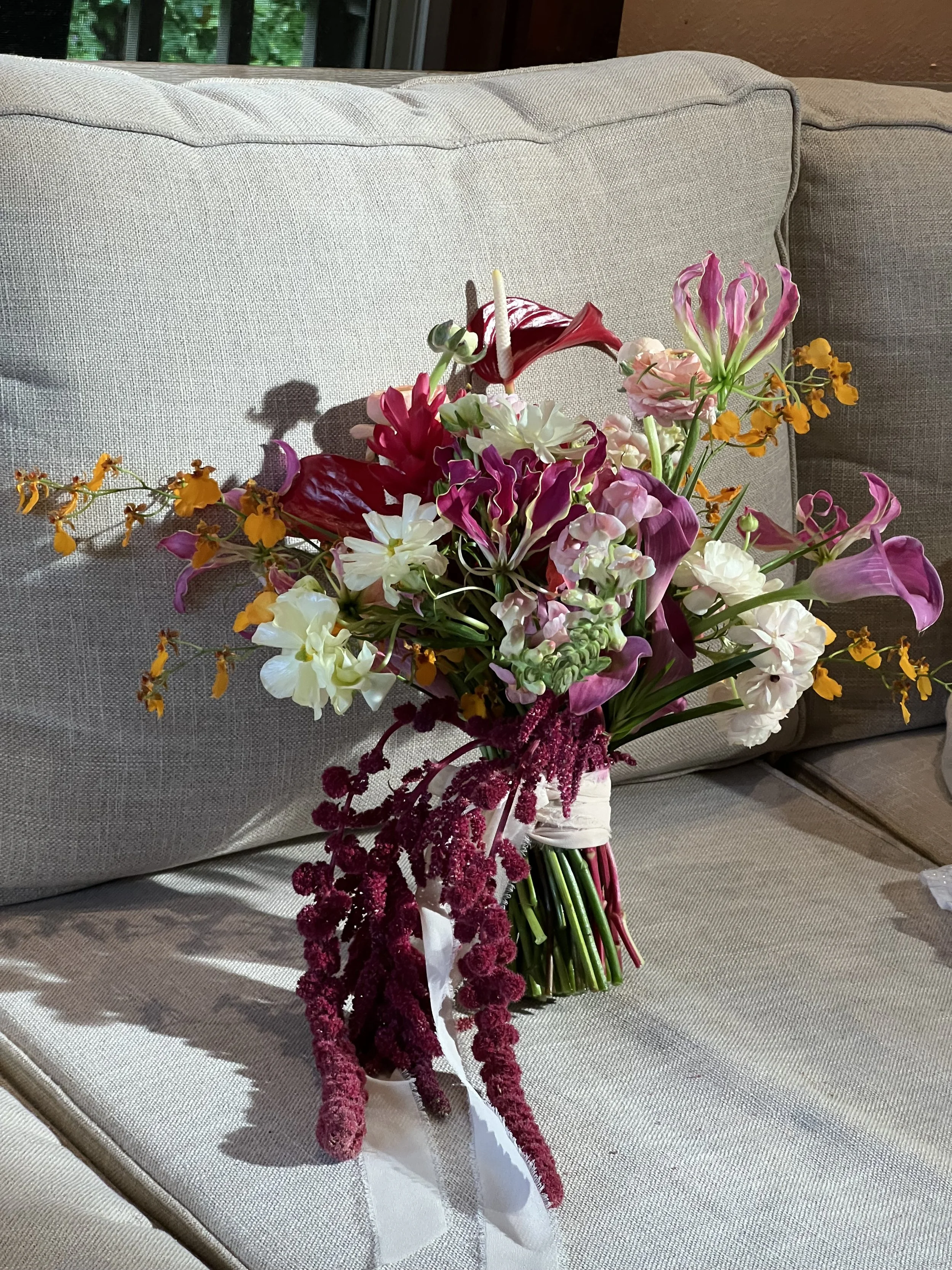 A colorful bouquet of various flowers, including calla lilies, roses, and other blossoms, resting on a light gray fabric sofa with a window in the background.