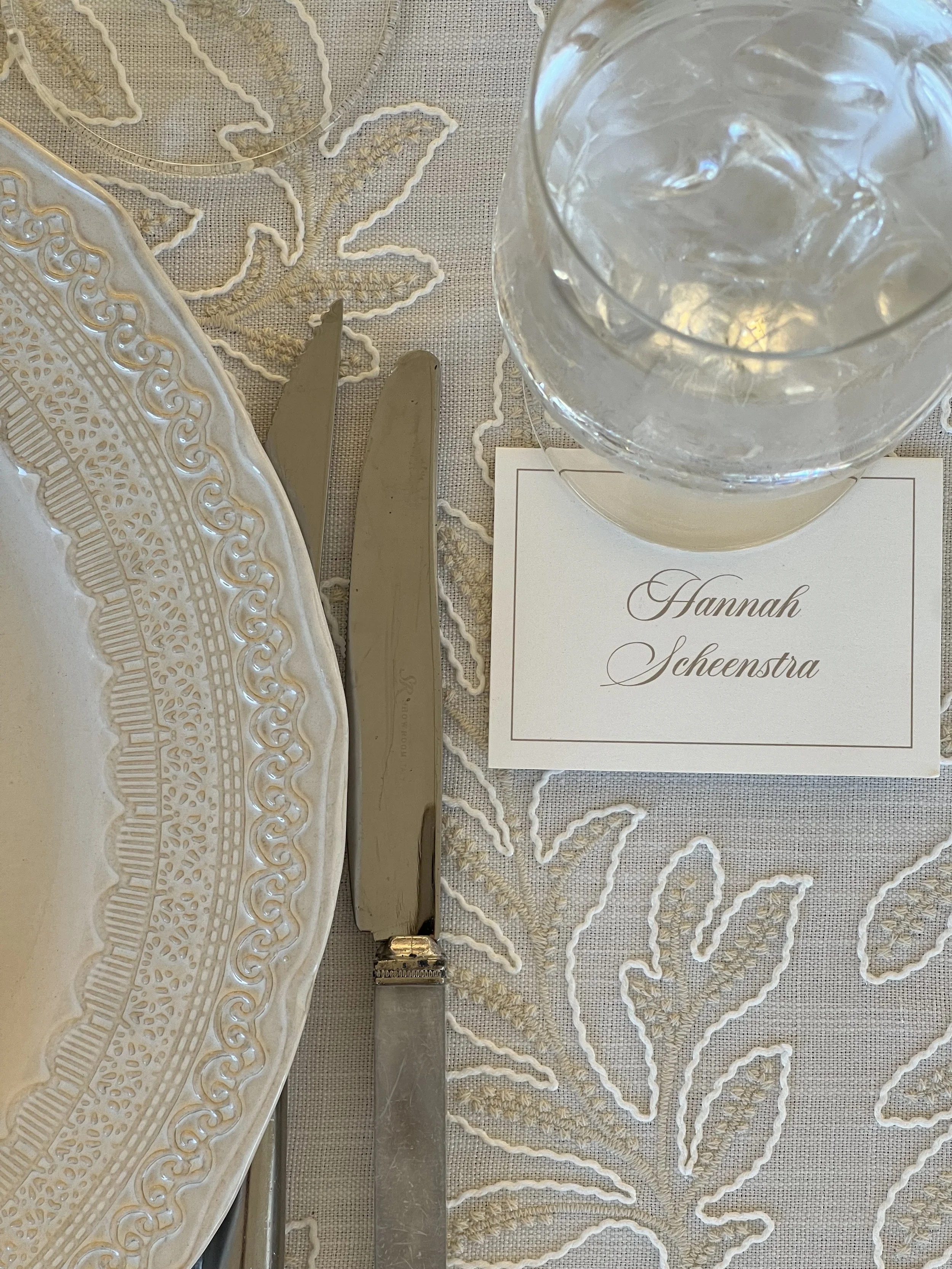 A table setting with a decorative oval white lace plate, a knife, a fork, and a place card with the name 'Hannah Scheintra.' There is also a glass of water and a napkin with embroidered floral patterns.