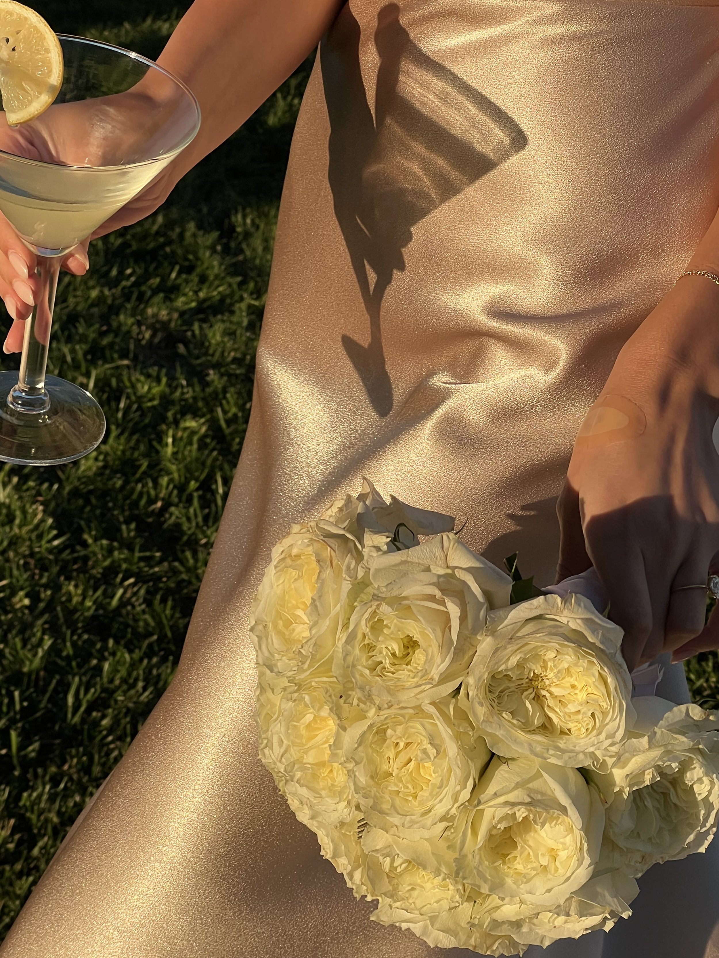 A person holding a coupe glass with a lemon wedge on the rim, a bouquet of cream-colored roses, and a gold shimmering fabric with a shadow of a glass cast on it. The setting appears to be outdoors in sunlight.