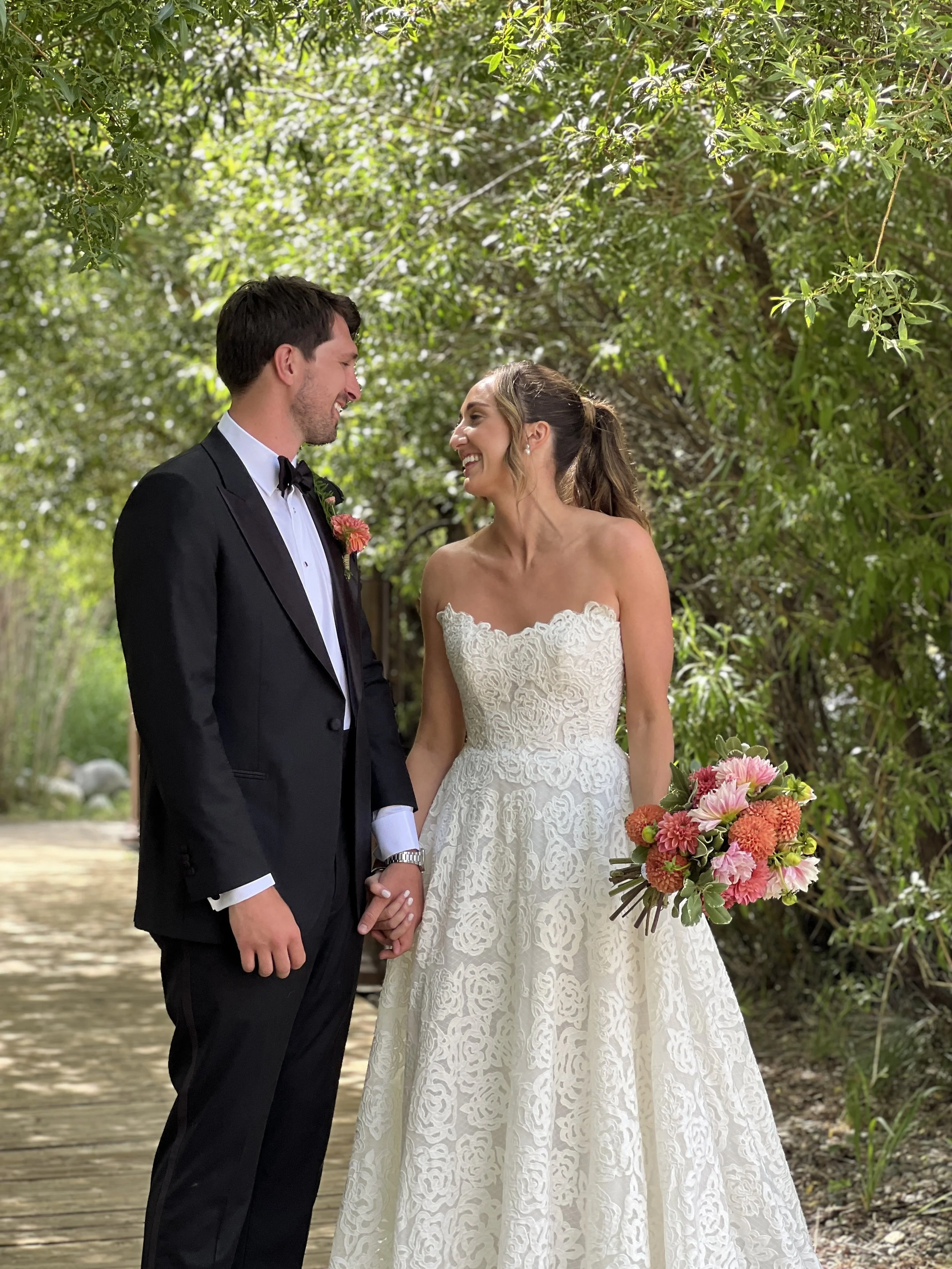A bride and groom standing outdoors, holding hands, smiling and looking at each other, surrounded by green foliage. The bride wears a strapless white lace wedding gown and holds a colorful bouquet. The groom is dressed in a black tuxedo with a bow ti